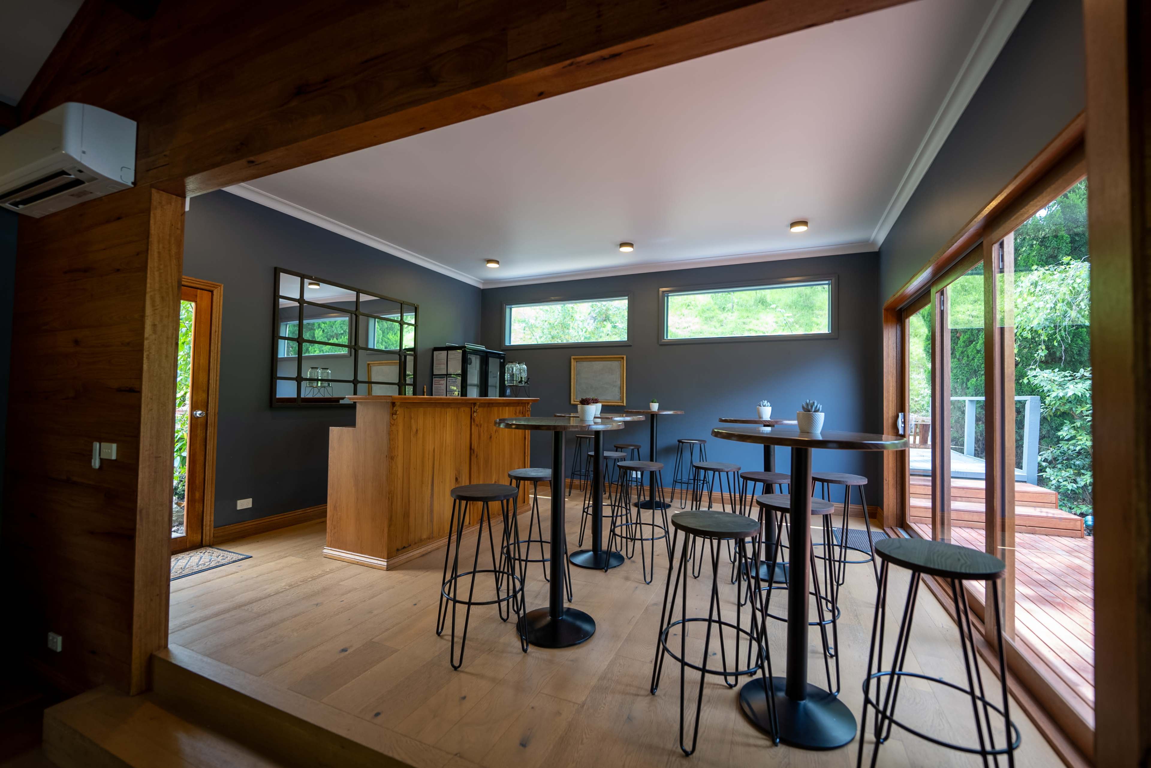 A modern interior space with a wooden bar, several high tables and stools, and large windows that open to a deck surrounded by greenery.