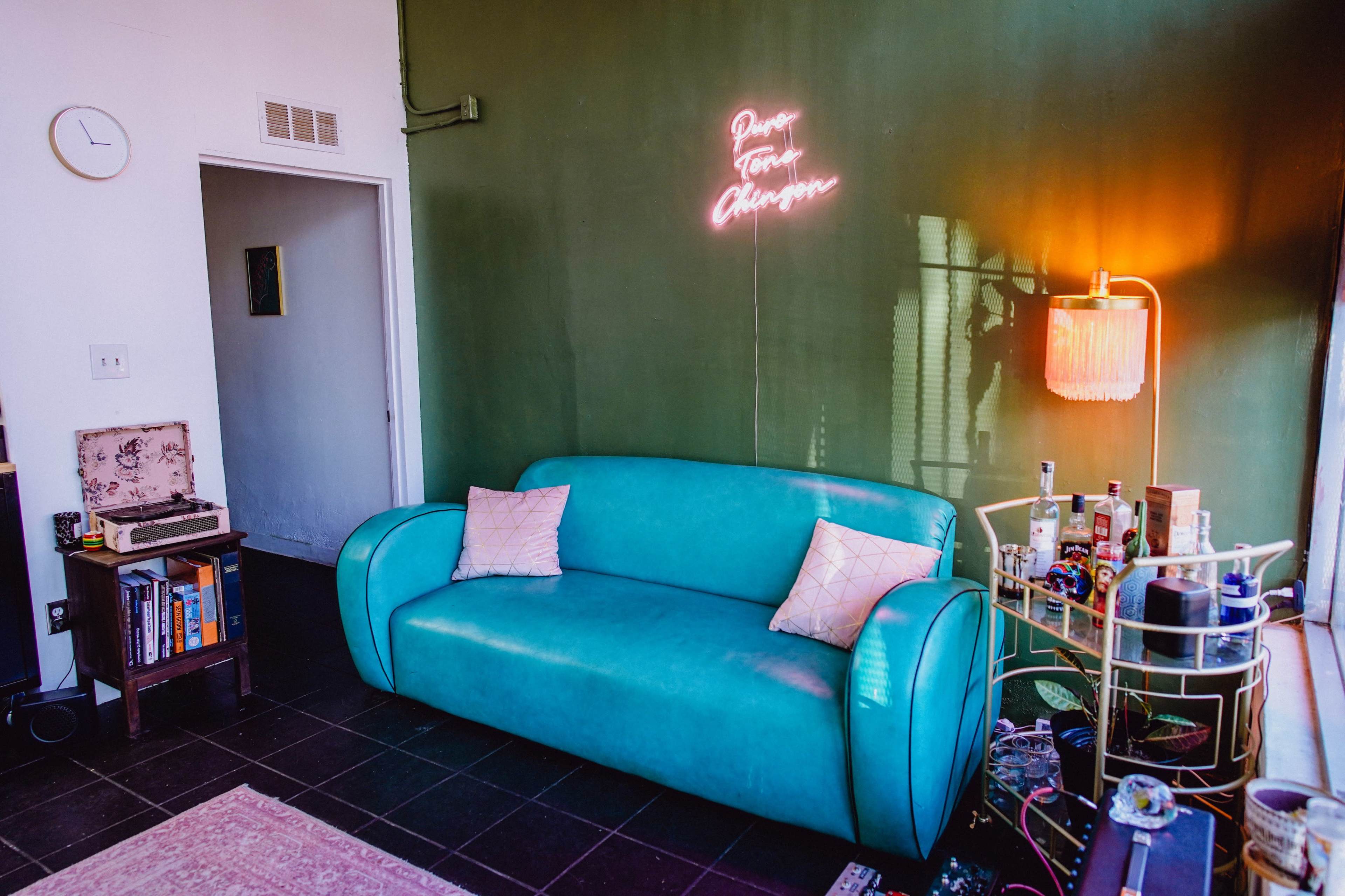 The image shows a bright turquoise couch against a green wall, accompanied by a neon sign, a lamp, and a collection of items on a small table.