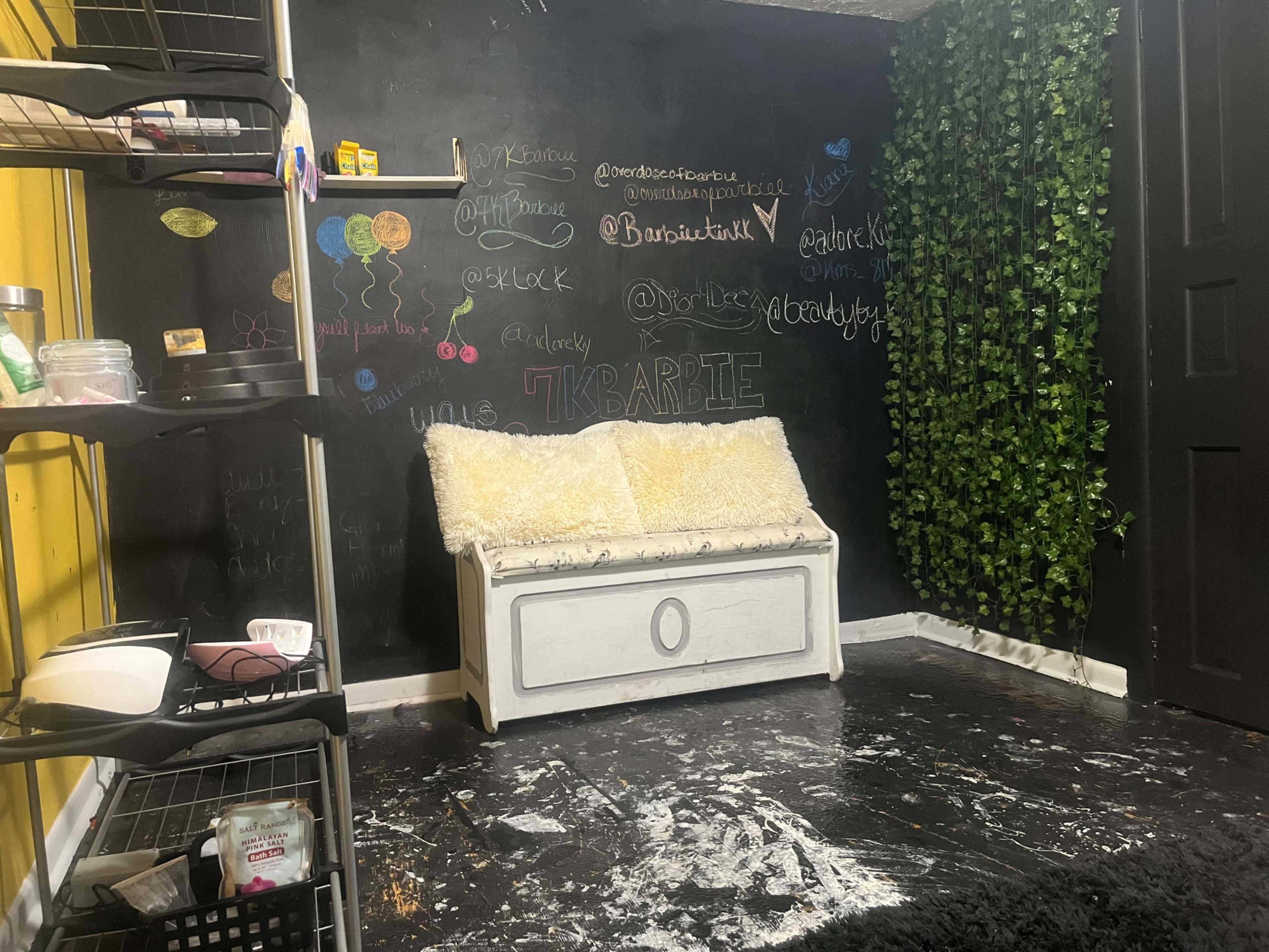A room featuring a black chalkboard wall with various colorful writings, a white storage bench with a fluffy cover, and a section of greenery alongside a black door.