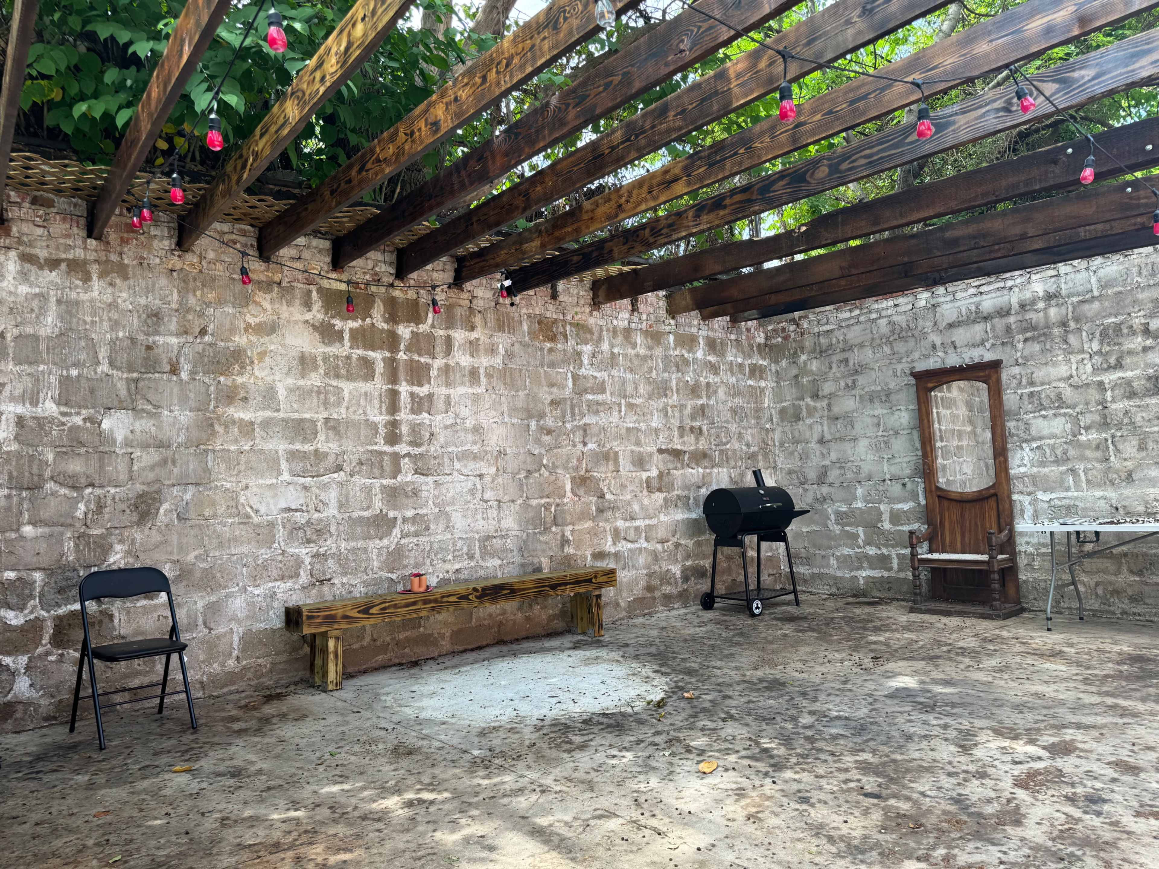 An empty outdoor space with stone walls, wooden beams overhead, a bench, a chair, a barbecue grill, and a table against the wall.