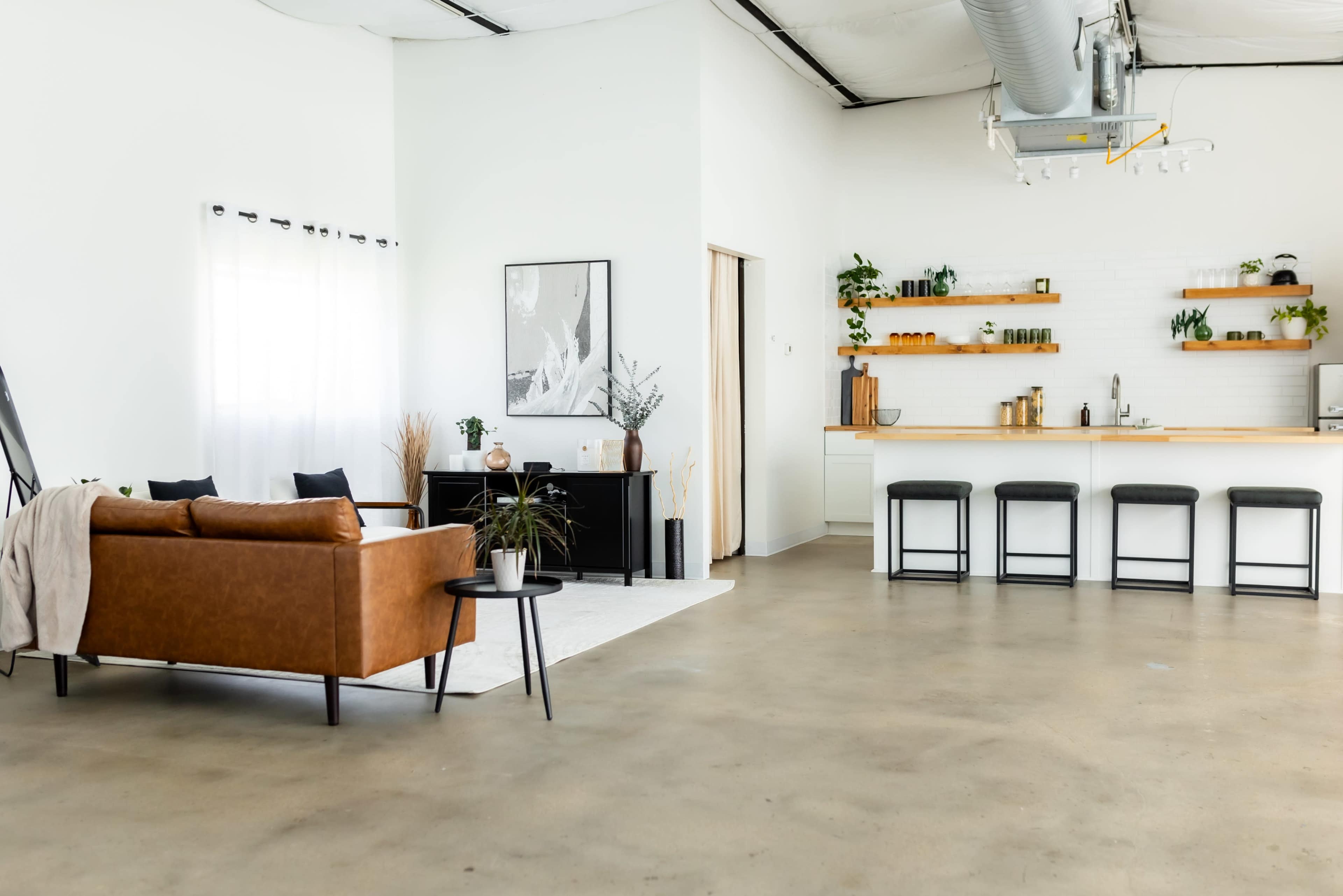 A modern, open-concept living space featuring a brown leather sofa, a black console table, and a kitchen area with wooden shelves and bar stools.