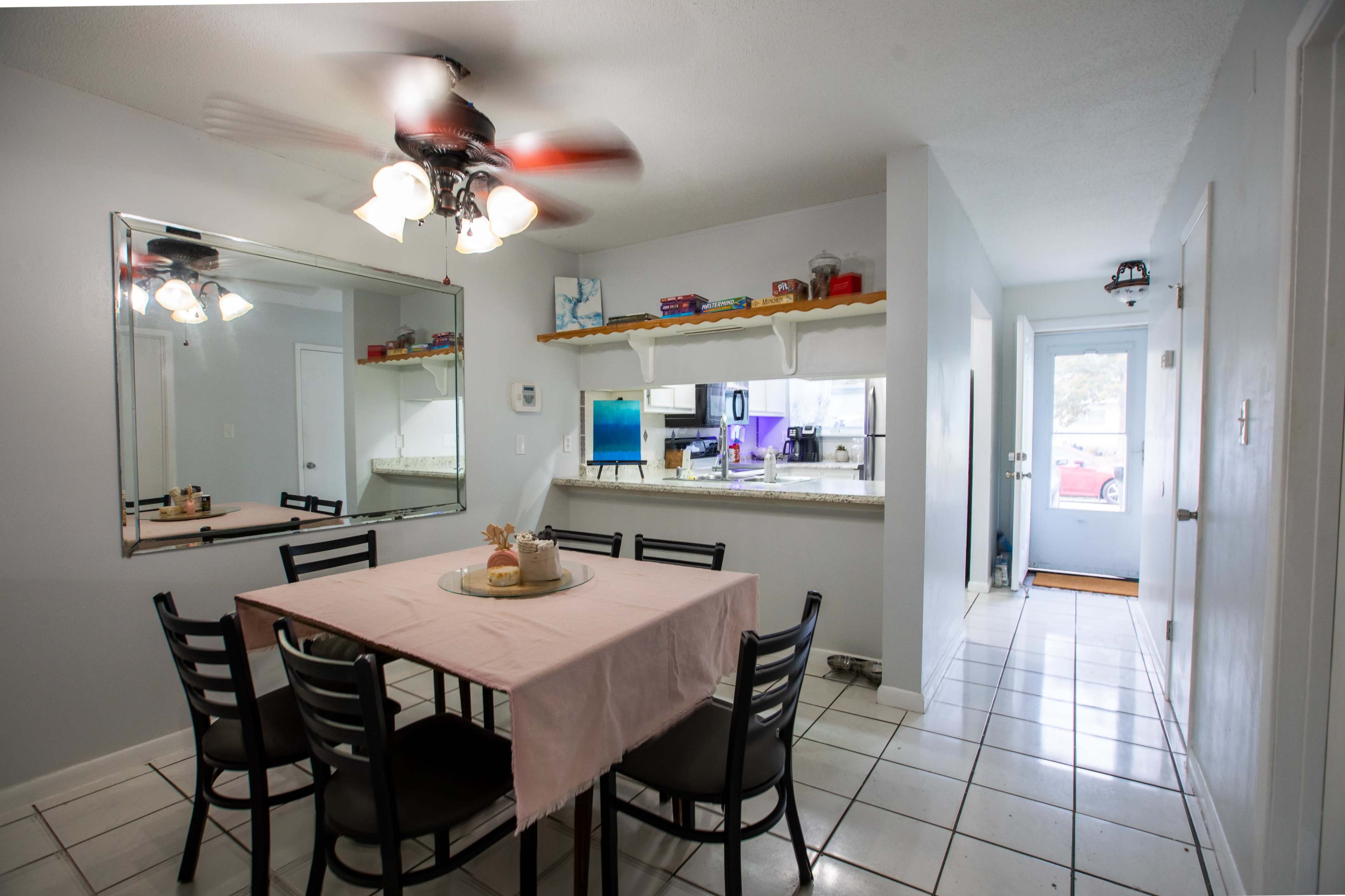 A dining area features a rectangular table with a pink tablecloth surrounded by black chairs, with a mirror and a view of a kitchen in the background.