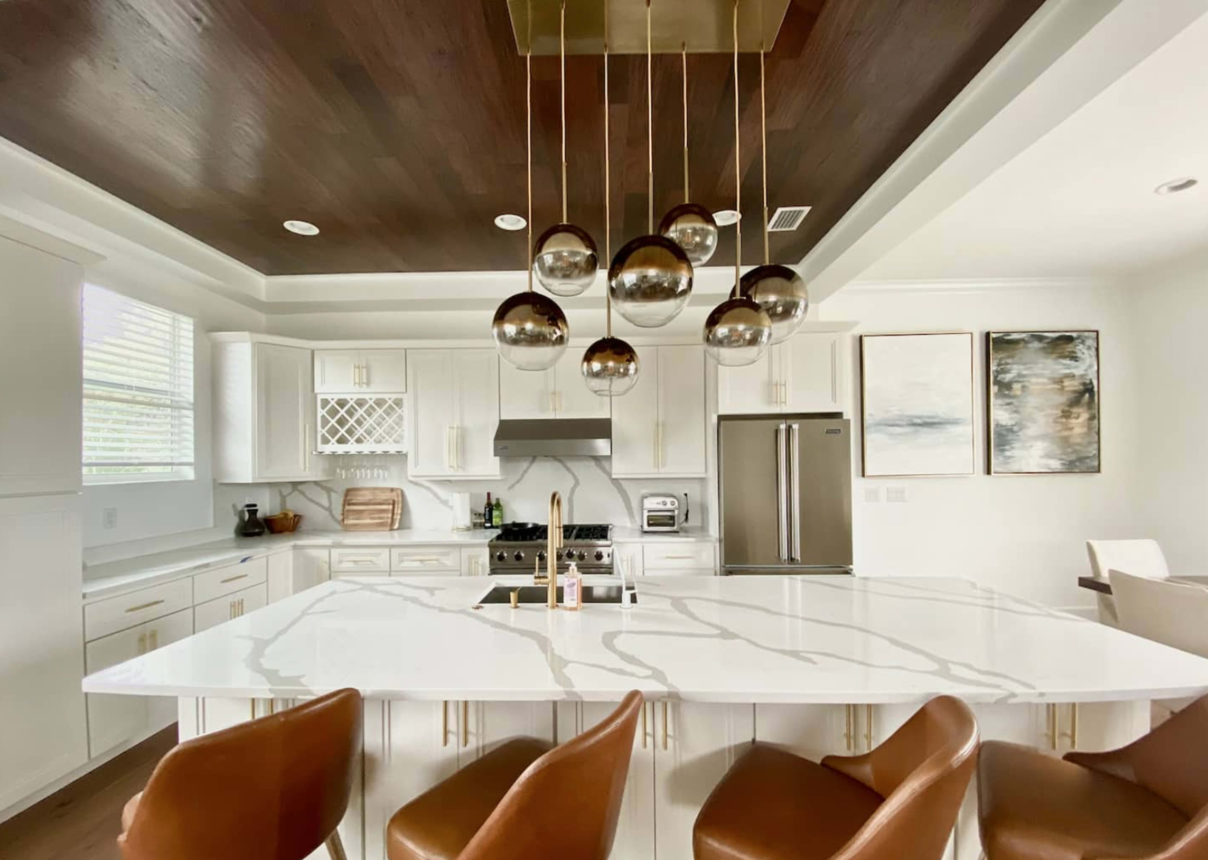 A modern kitchen features a large marble island with metal bar stools, pendant lighting, and stainless steel appliances.