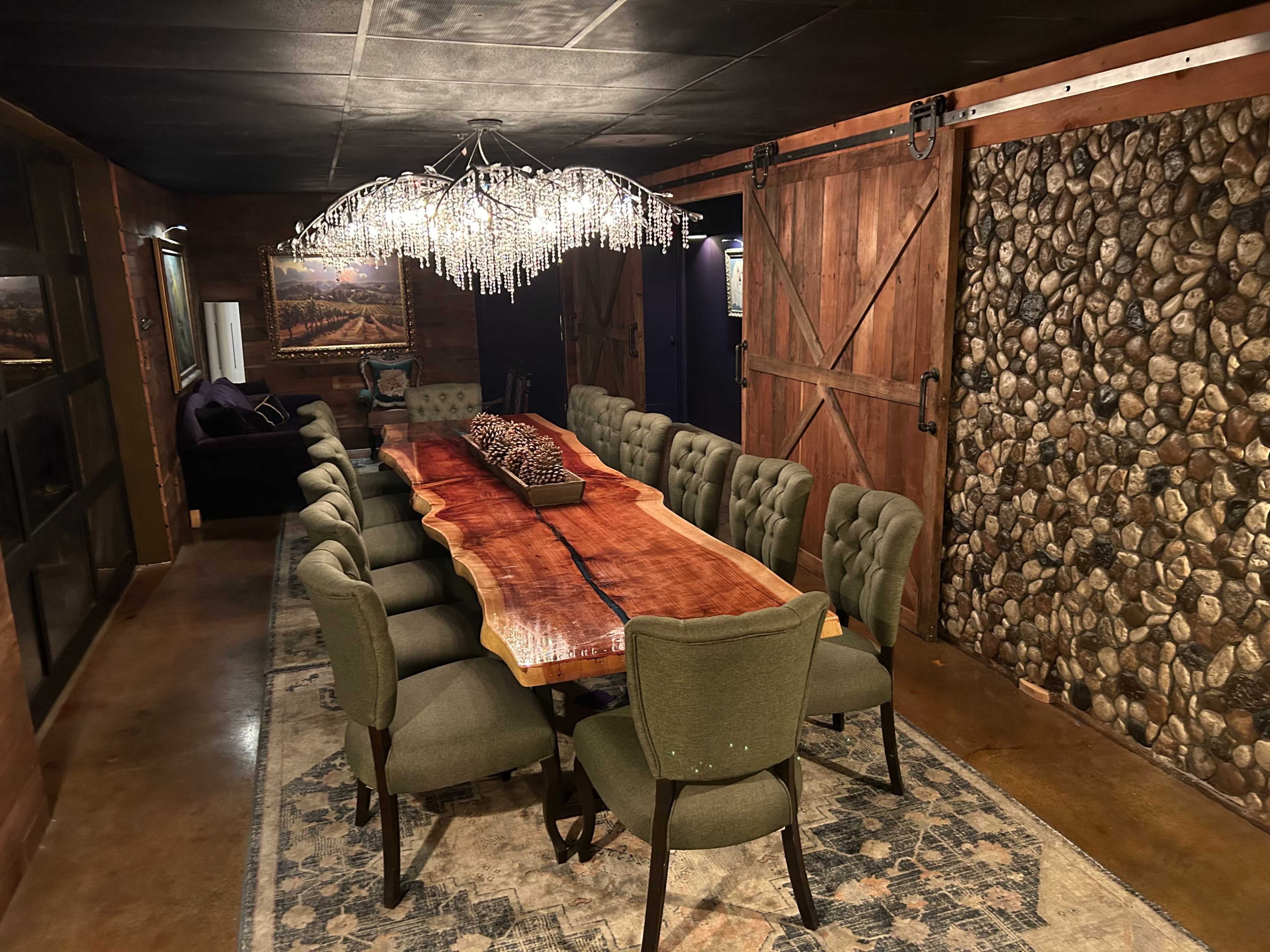 A large wooden dining table surrounded by ten green upholstered chairs in a room with rustic wooden walls and a chandelier made of crystals.