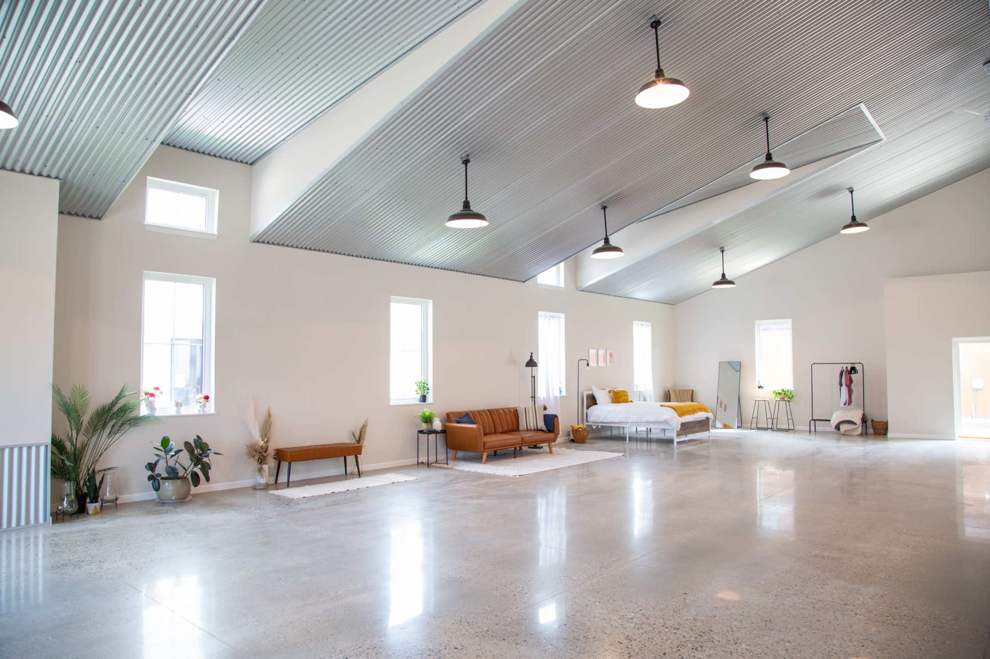 The image shows a spacious room with high ceilings, featuring a mix of seating, a bed, and large windows, illuminated by pendant lights.