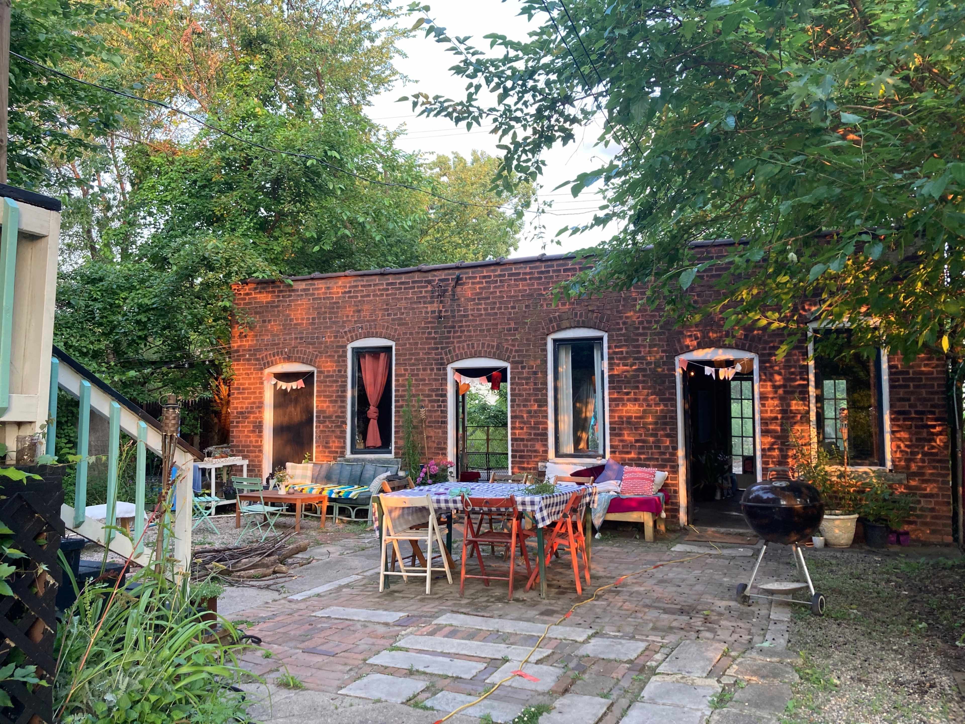 The scene shows a brick building with multiple windows, surrounded by trees, featuring an outdoor seating area and a barbecue grill.