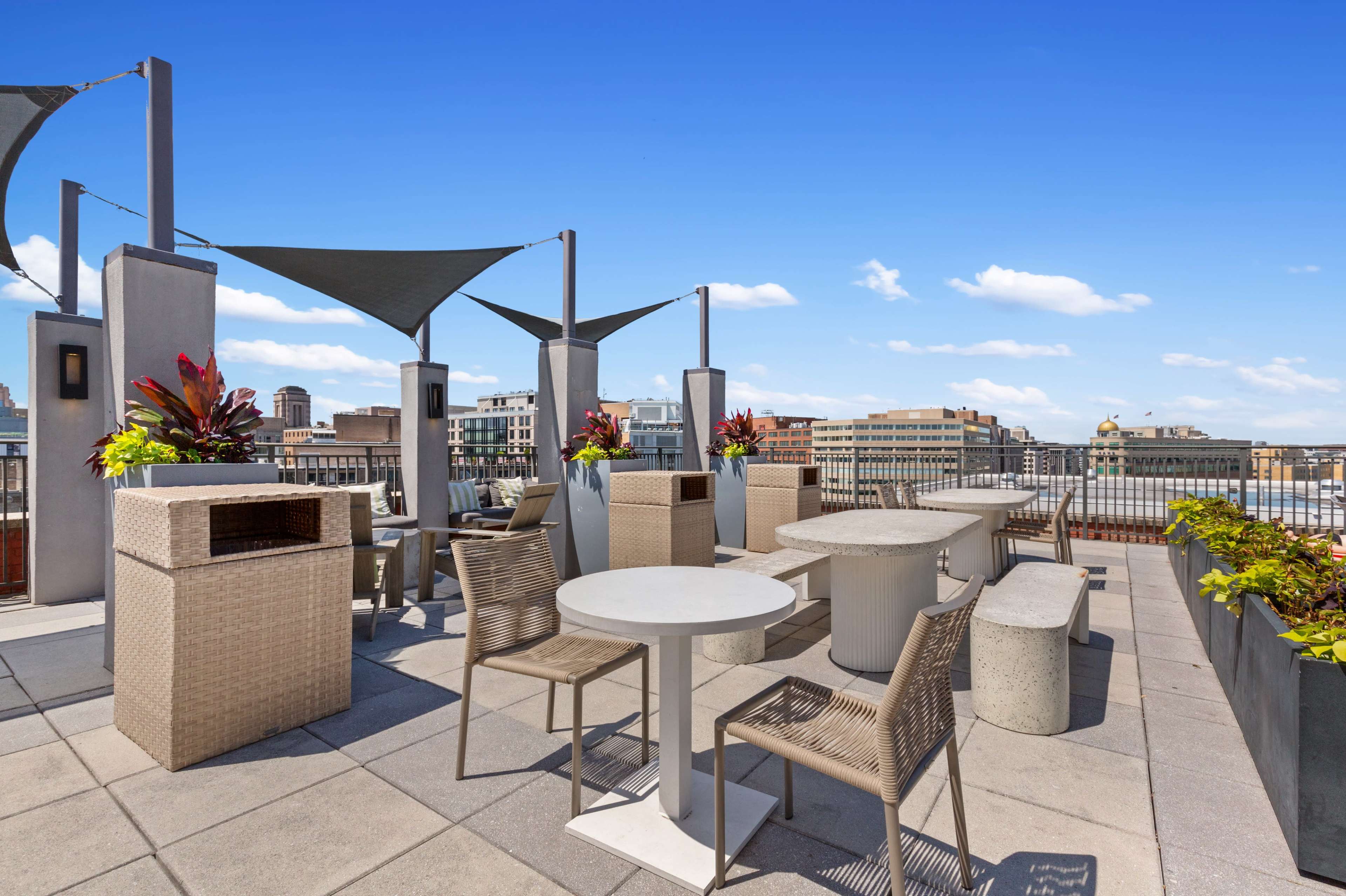The image shows a rooftop terrace with outdoor furniture, including tables and chairs, surrounded by planters and shade sails against a clear blue sky.