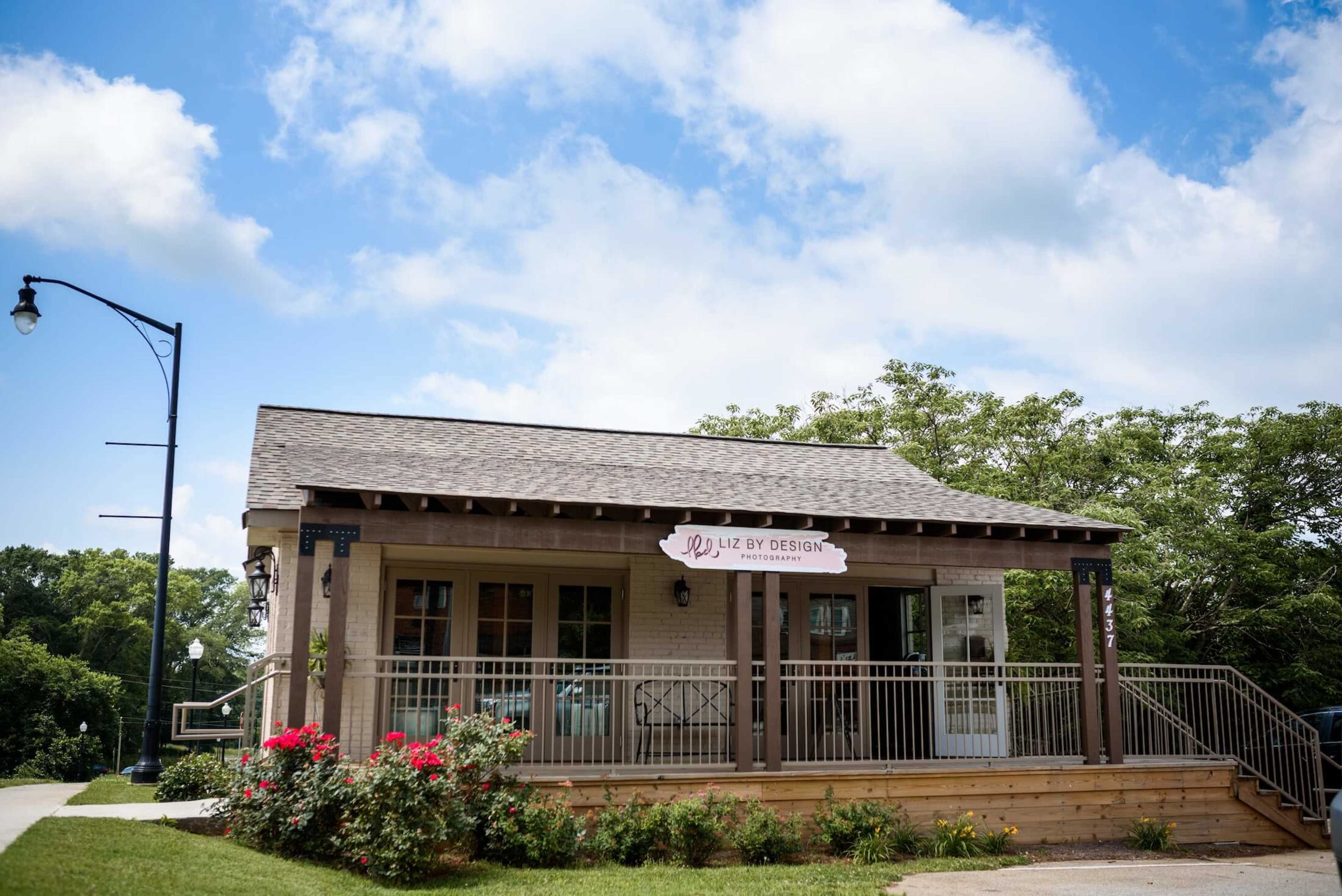 The image shows a small, single-story commercial building with a sign reading "Luz by Design," surrounded by green trees and blooming flowers.