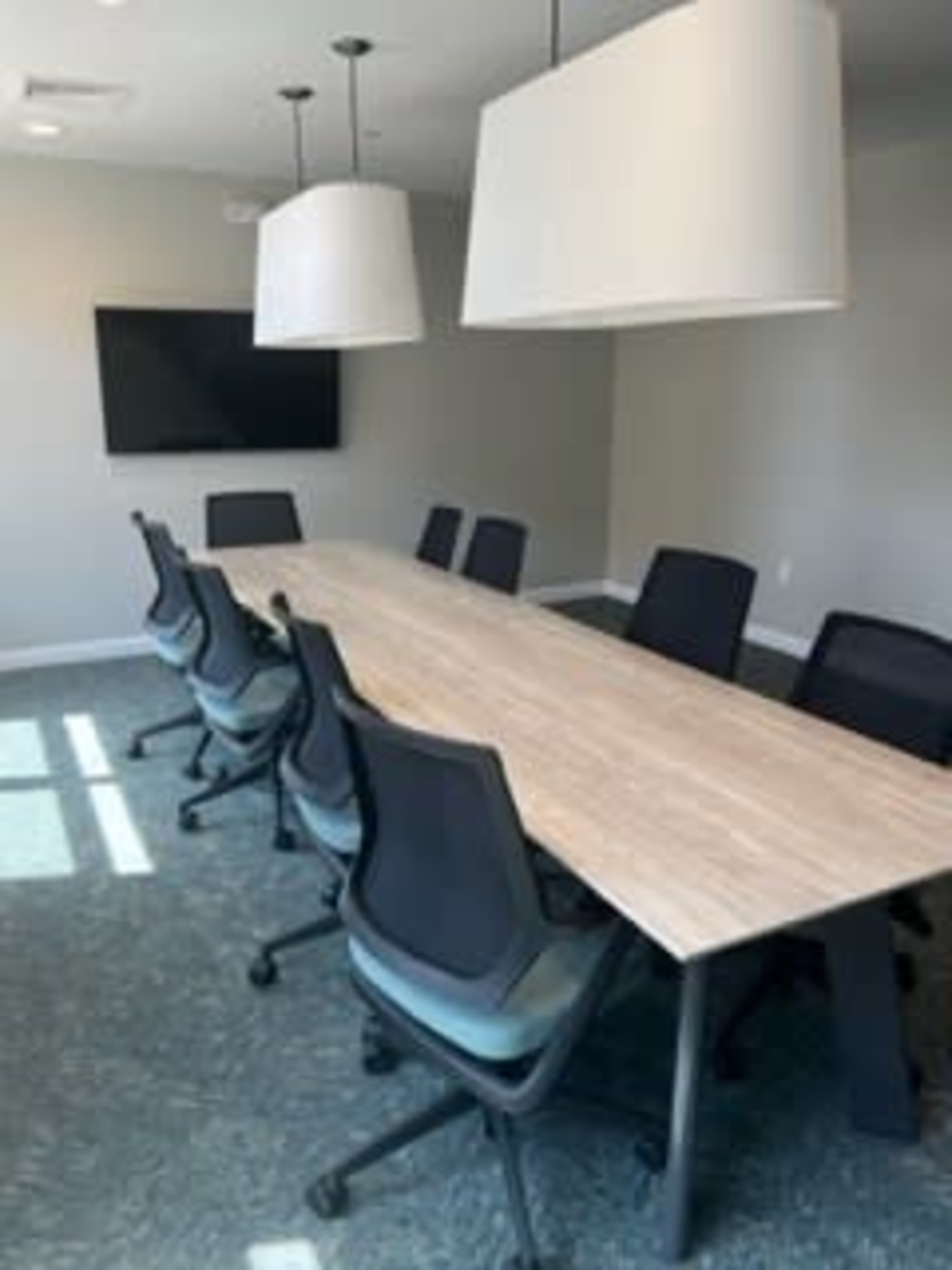 A modern conference room features a long wooden table surrounded by black rolling chairs and two large pendant lights above.