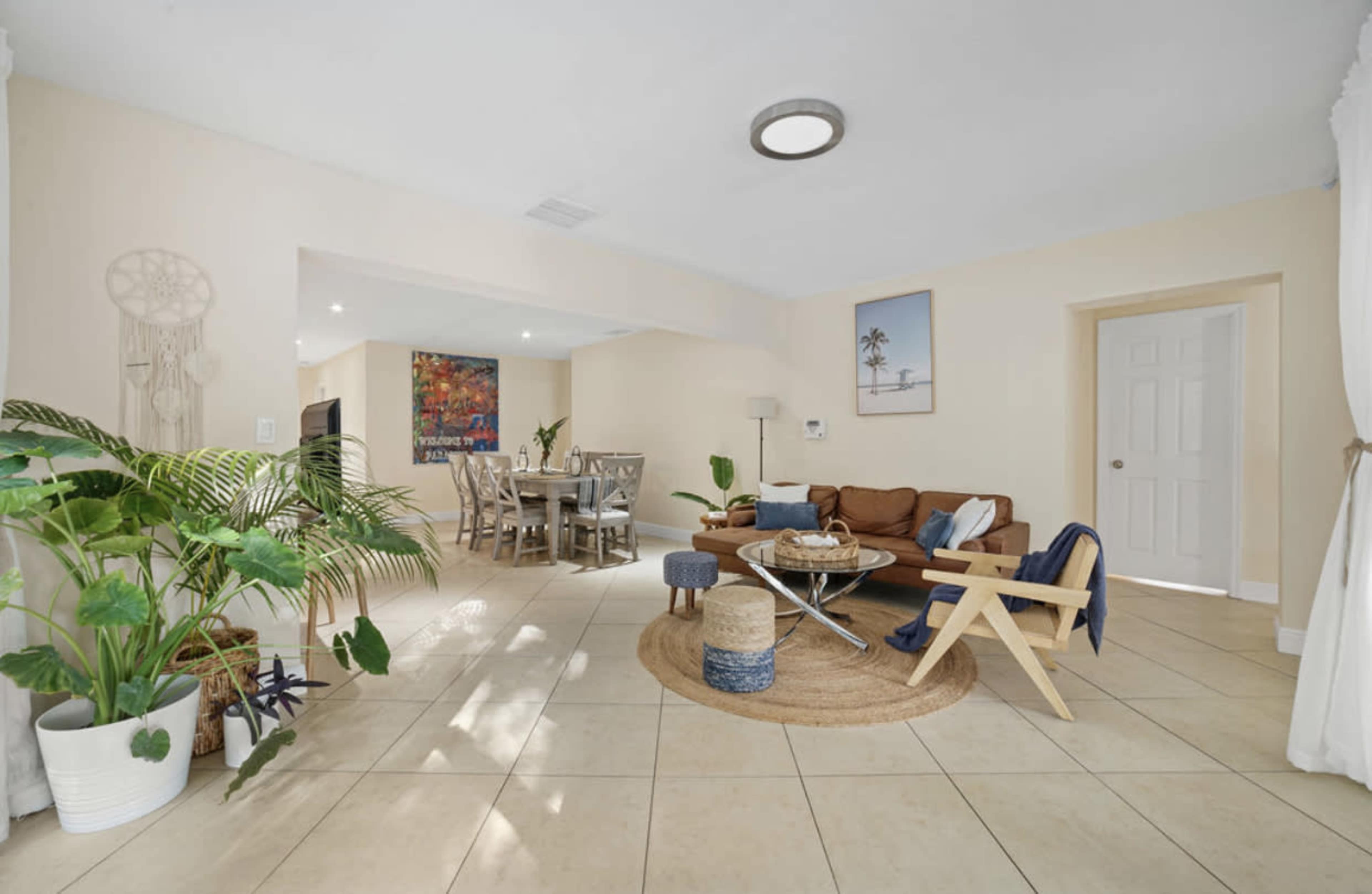 A bright living area features a sofa, coffee table, and plants, with a dining table visible in the background.