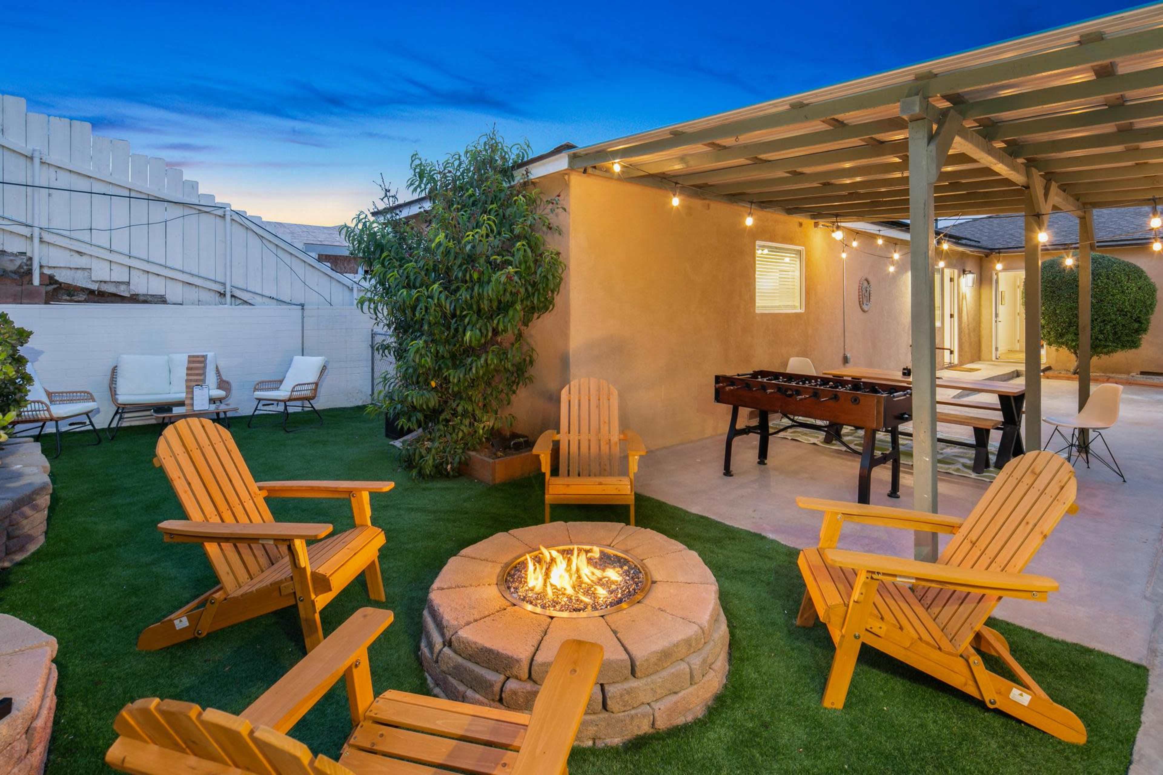 A fire pit surrounded by wooden chairs is situated in a backyard with a foosball table and string lights overhead.