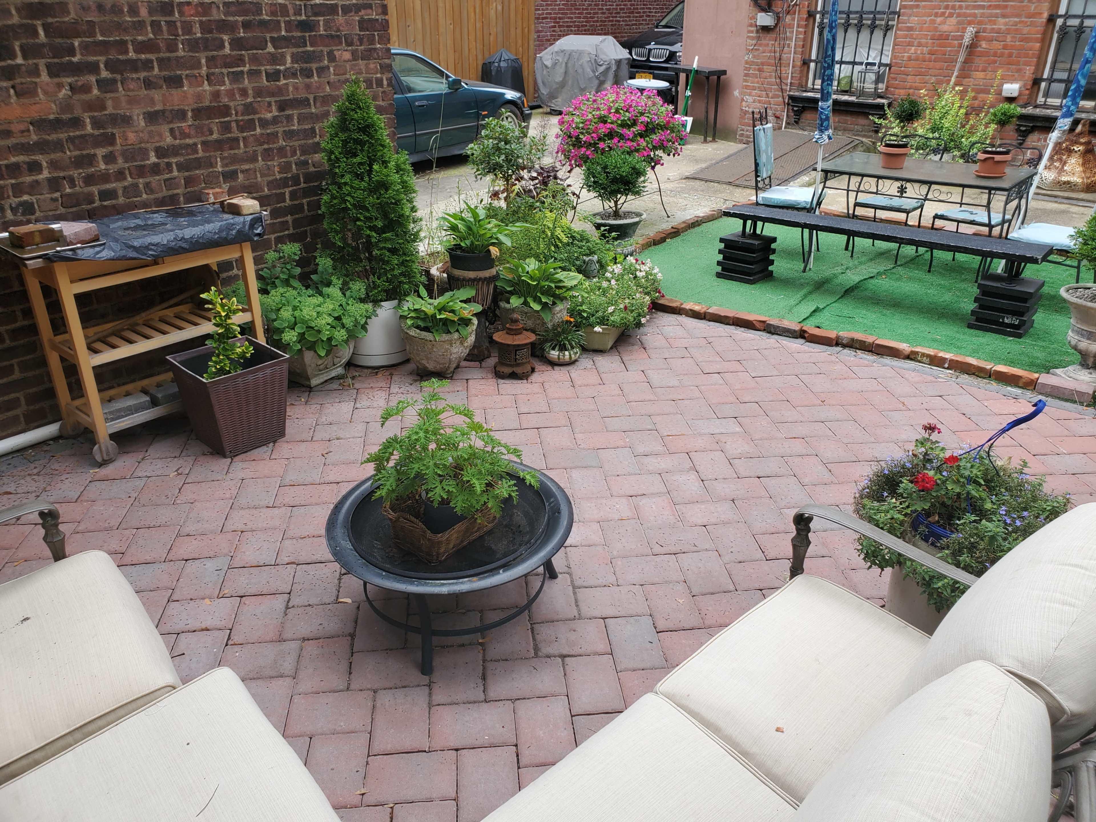 A brick-paved outdoor area features various potted plants, a small table, and seating arranged around a central fire pit.