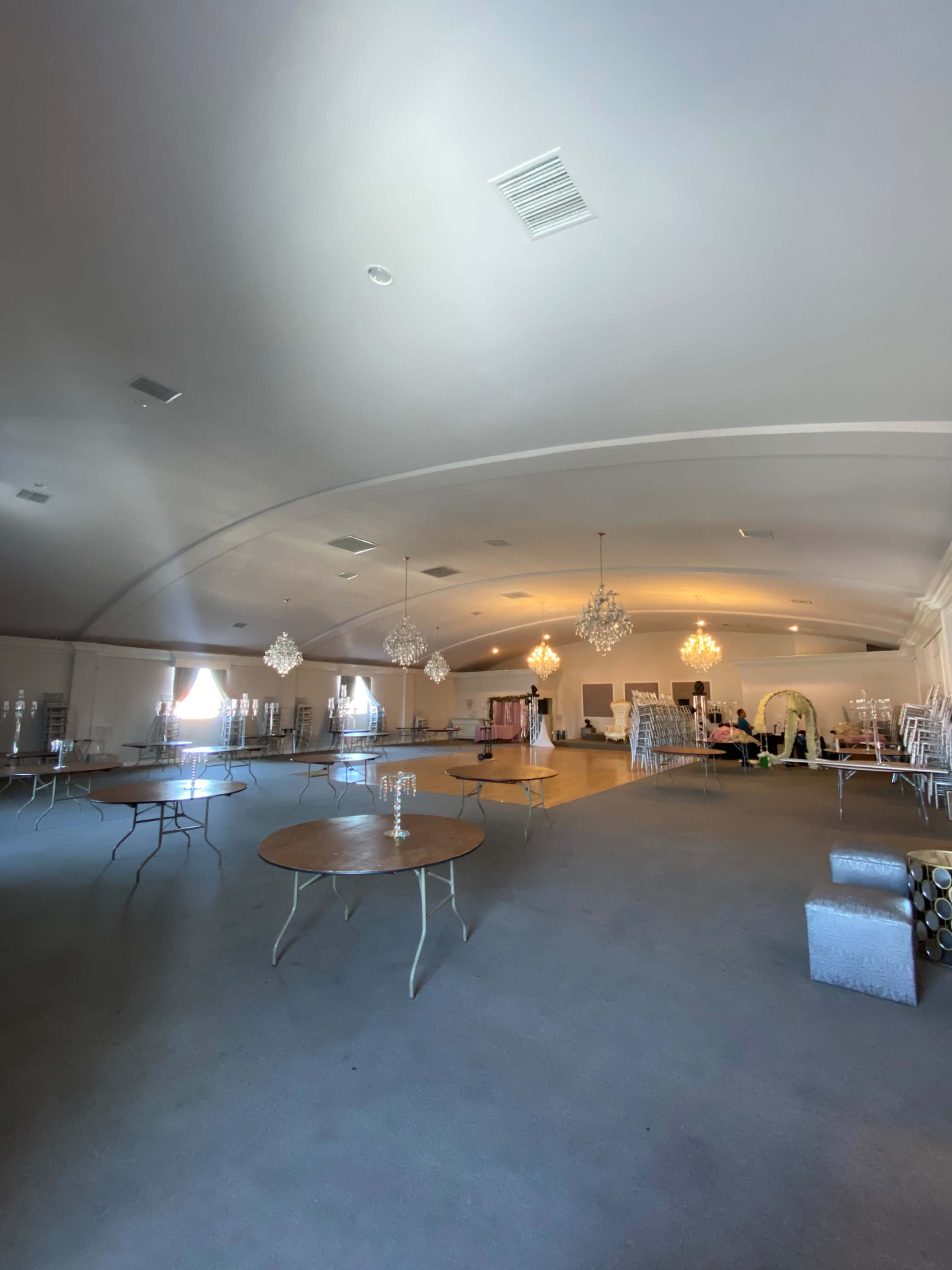A spacious, empty event hall with several round tables set up and chandeliers hanging from the ceiling.