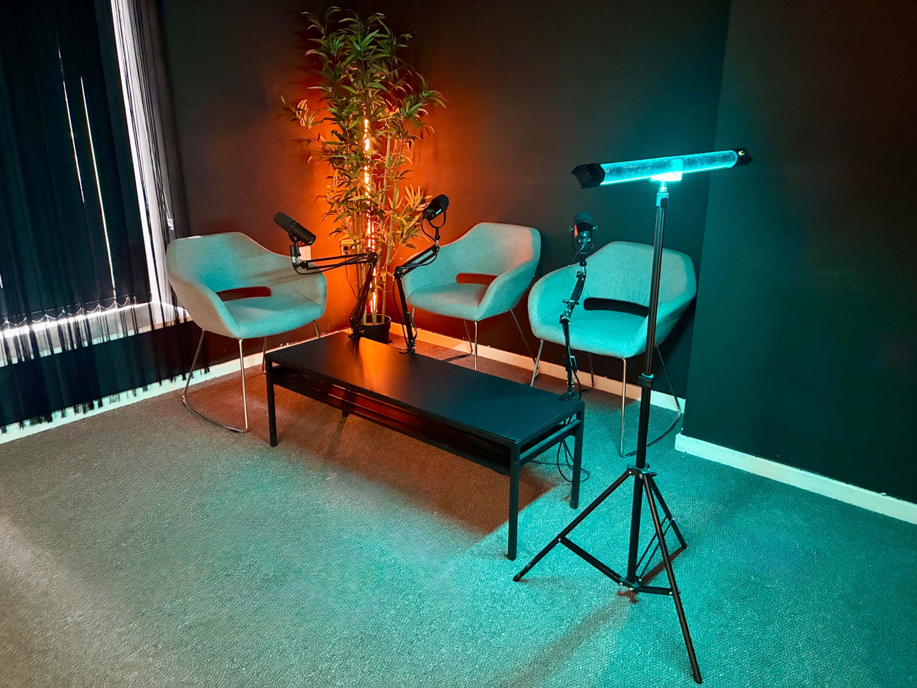 The image shows a small recording studio setup with two gray chairs, a table, and various lighting equipment against a dark wall.