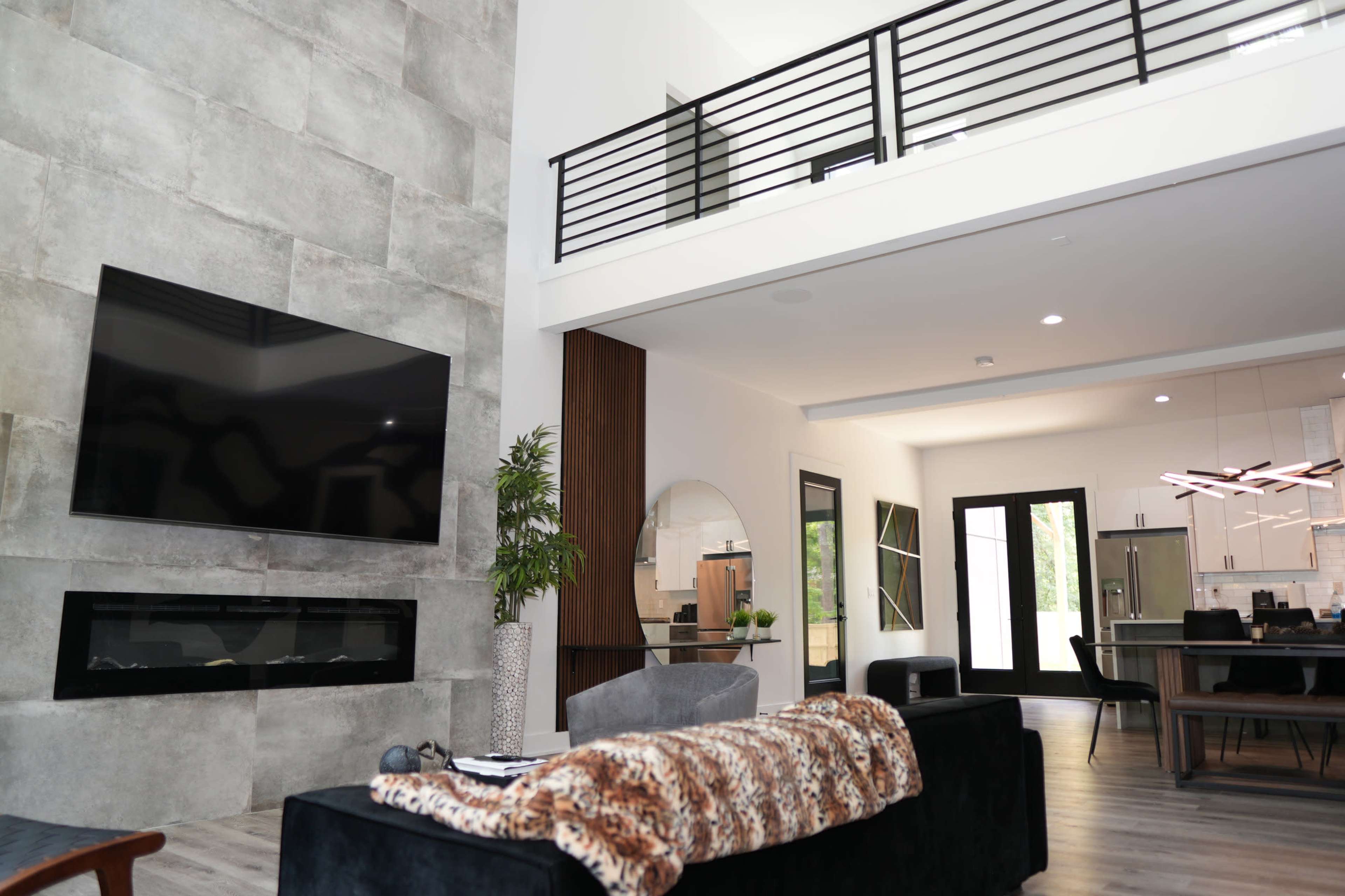 A modern living room with a large wall-mounted TV, a cozy sofa covered with a throw, and a sleek open kitchen visible through glass doors.
