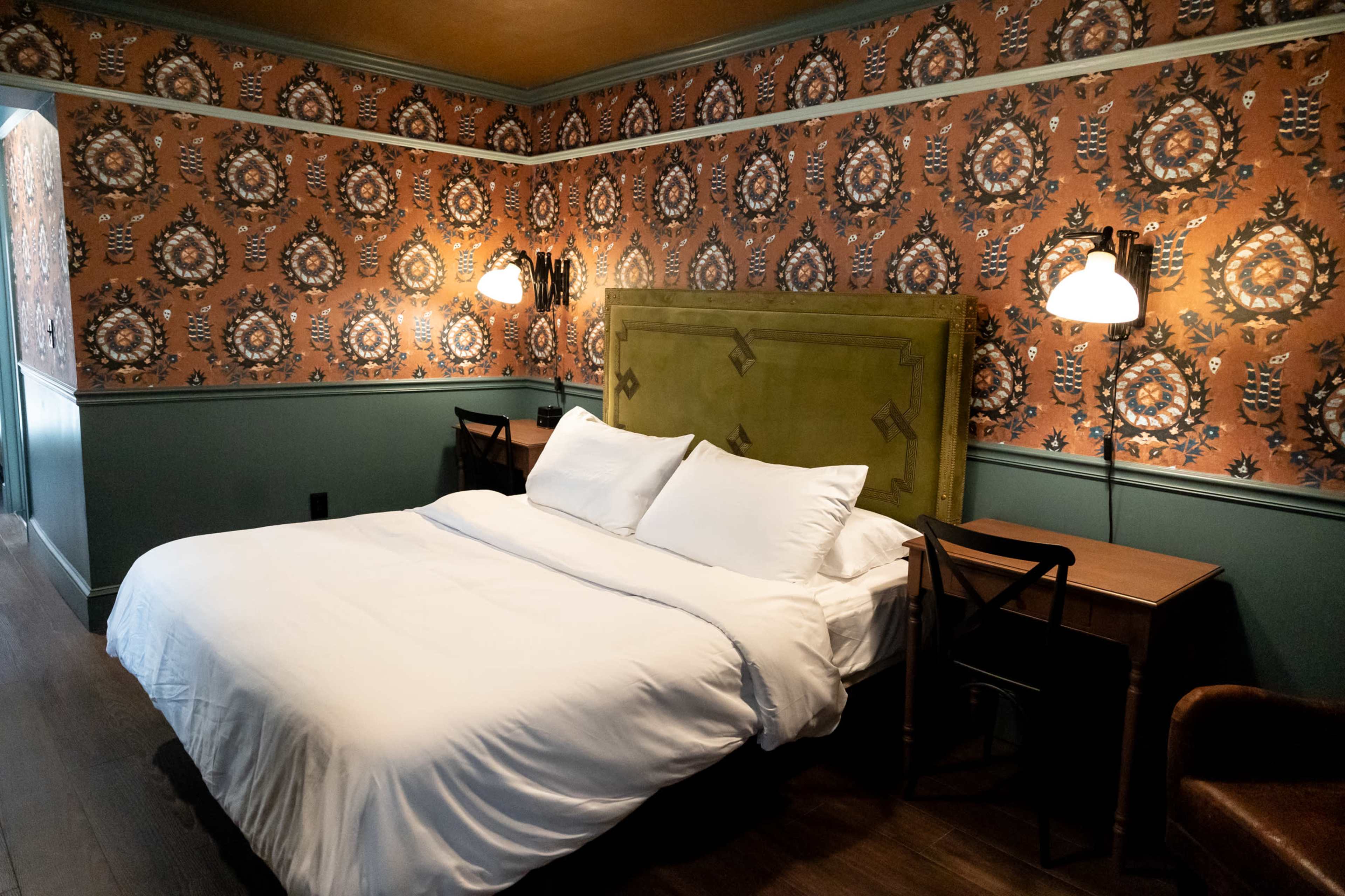A bedroom features a large bed with white bedding, a patterned wallpaper, and a small desk with chairs.