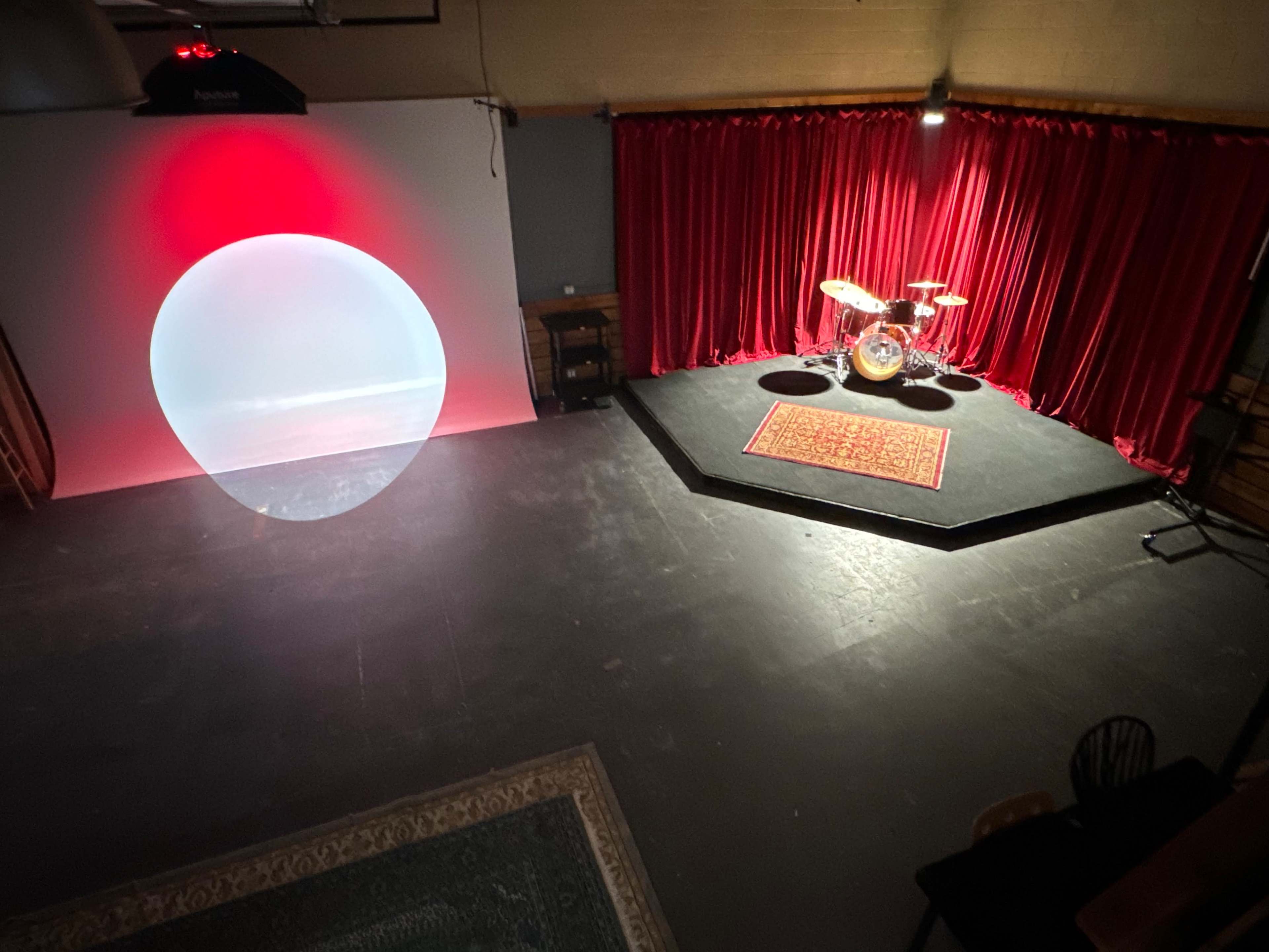 The image shows a staged performance area featuring a drum set on a raised platform surrounded by red curtains and a large circular light projection on the floor.