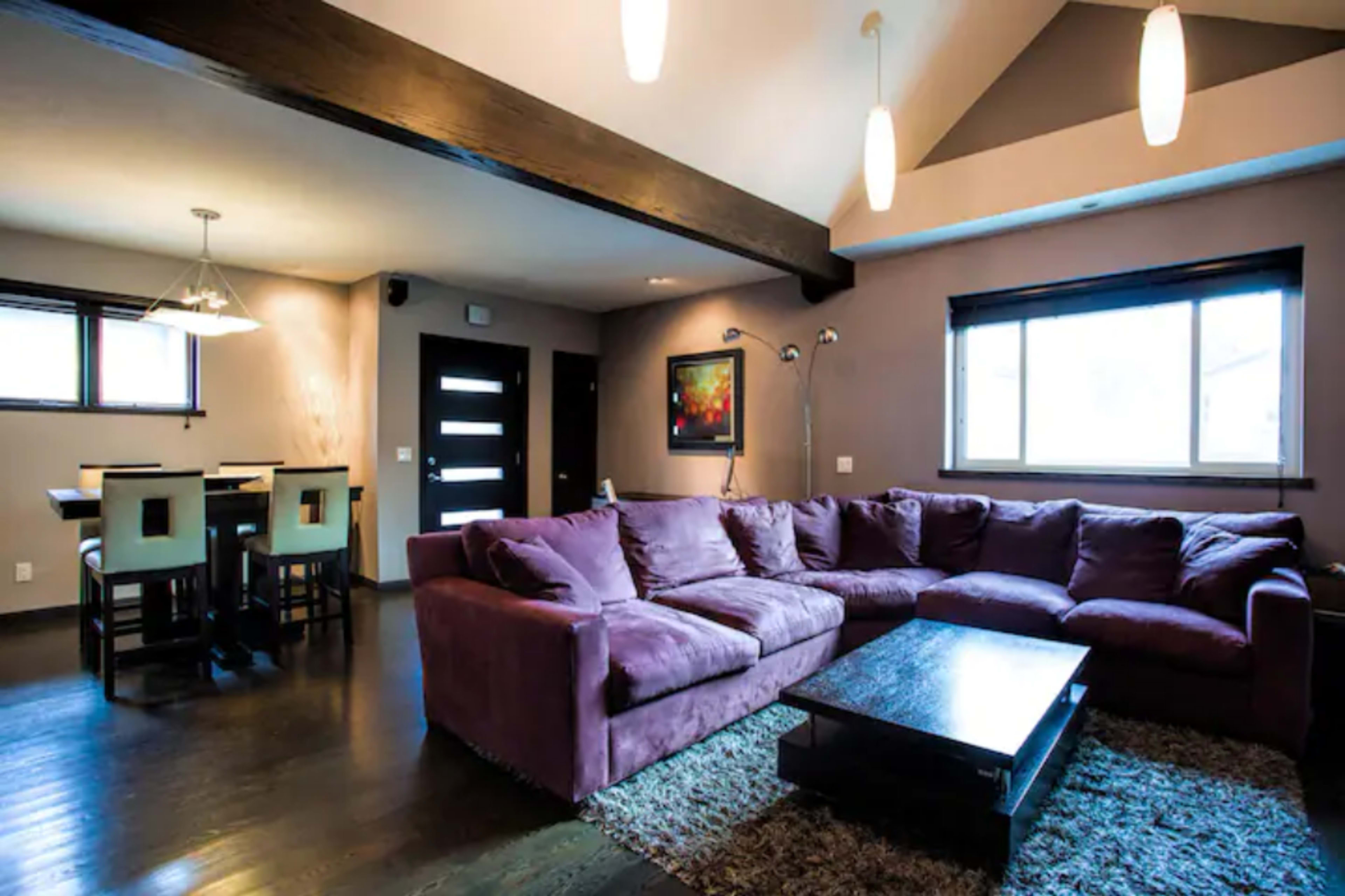 The image shows a modern living room featuring a large purple sectional sofa, a dark wooden coffee table, and a dining area with a table and chairs in the background.