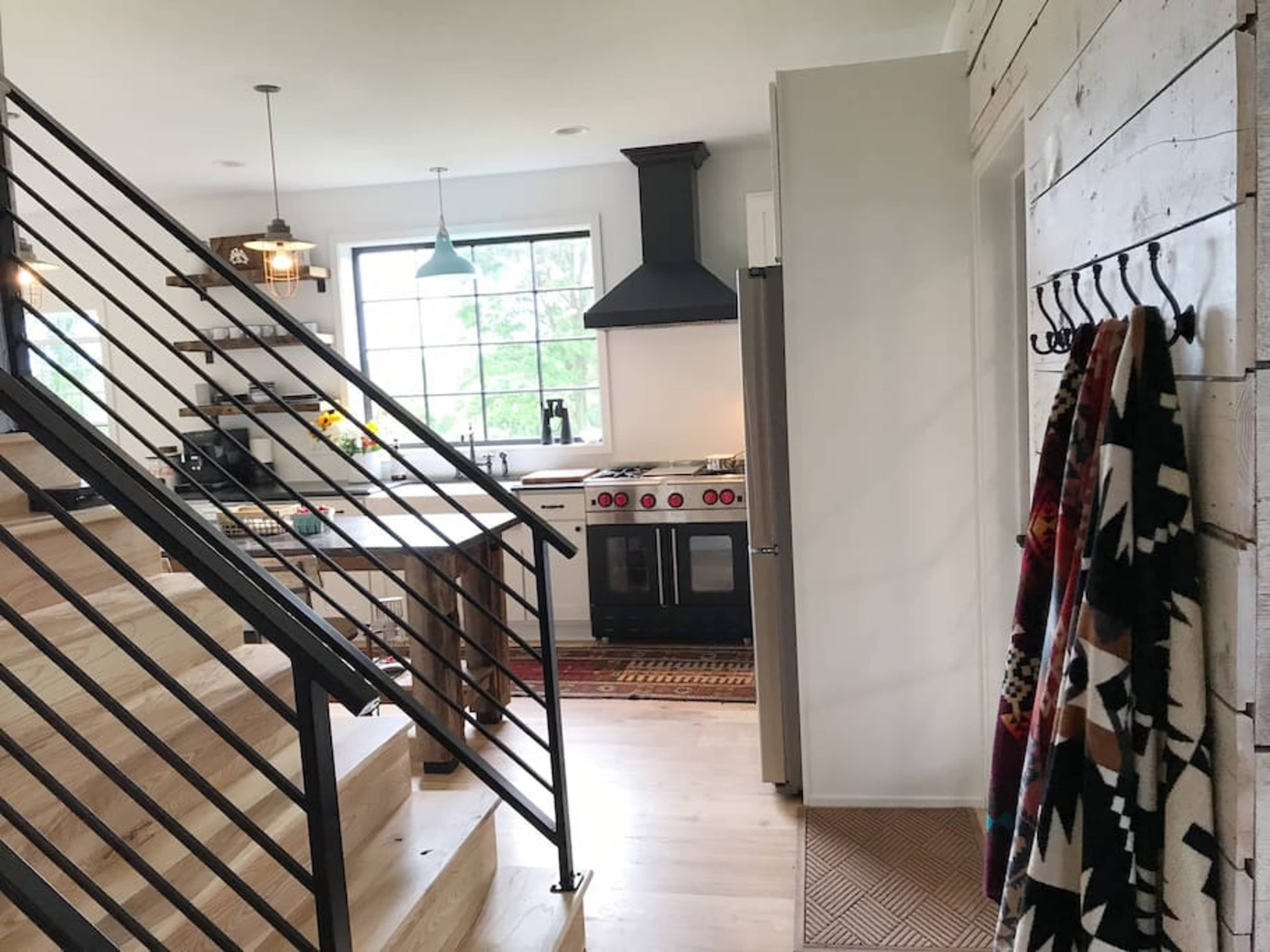 A modern kitchen is visible from a staircase, featuring a large window, a stove with red knobs, and wood accents.