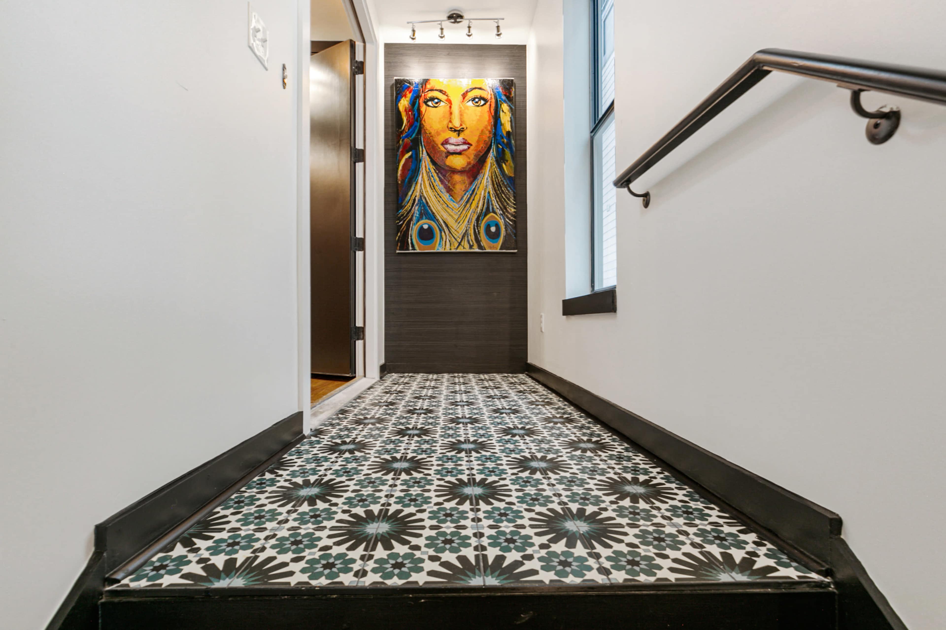 The image shows a hallway with patterned tile flooring leading to a dark door, illuminated by a brightly colored portrait on the wall.