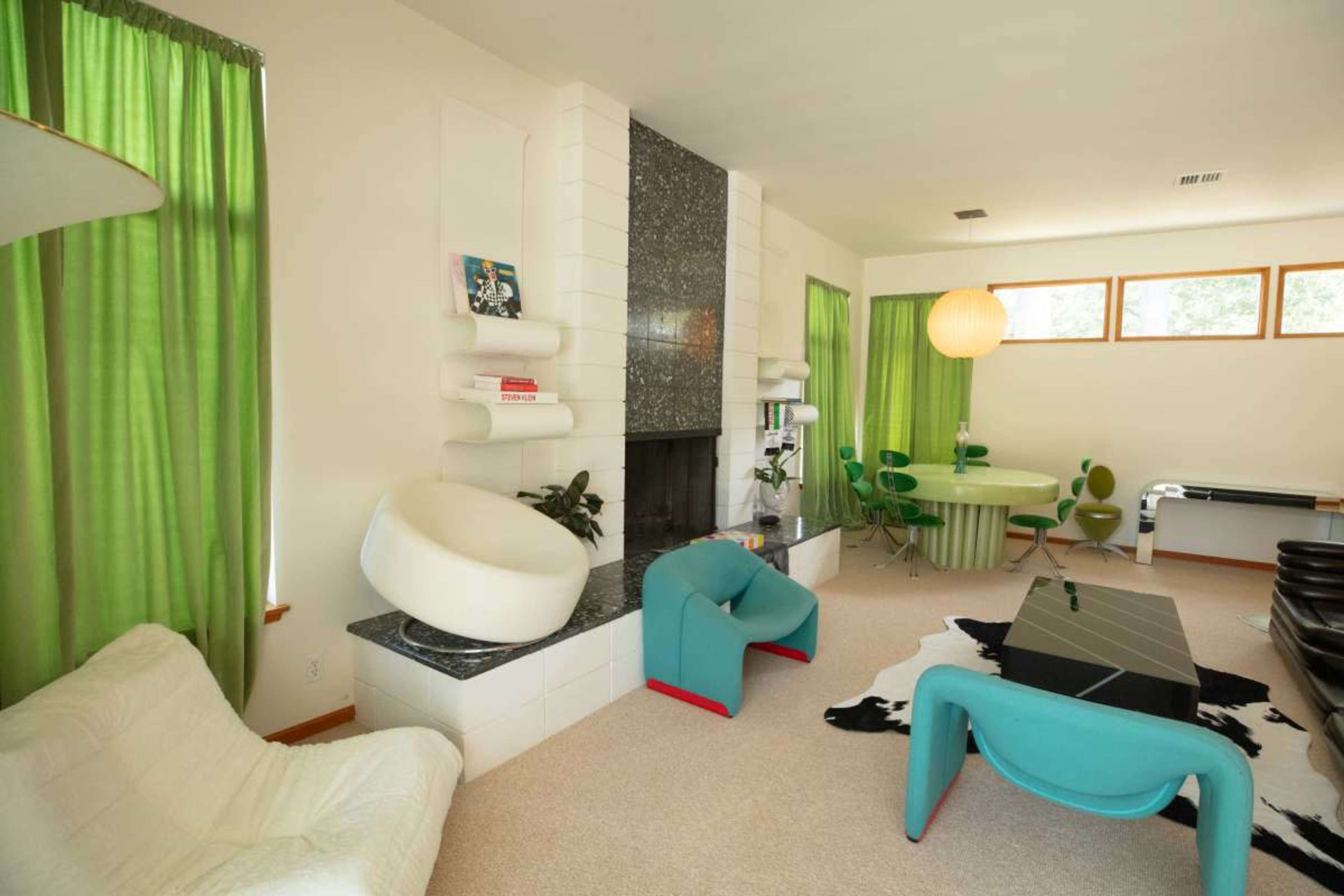 The image shows a modern living room featuring a fireplace, colorful furniture, green curtains, and a circular dining area.