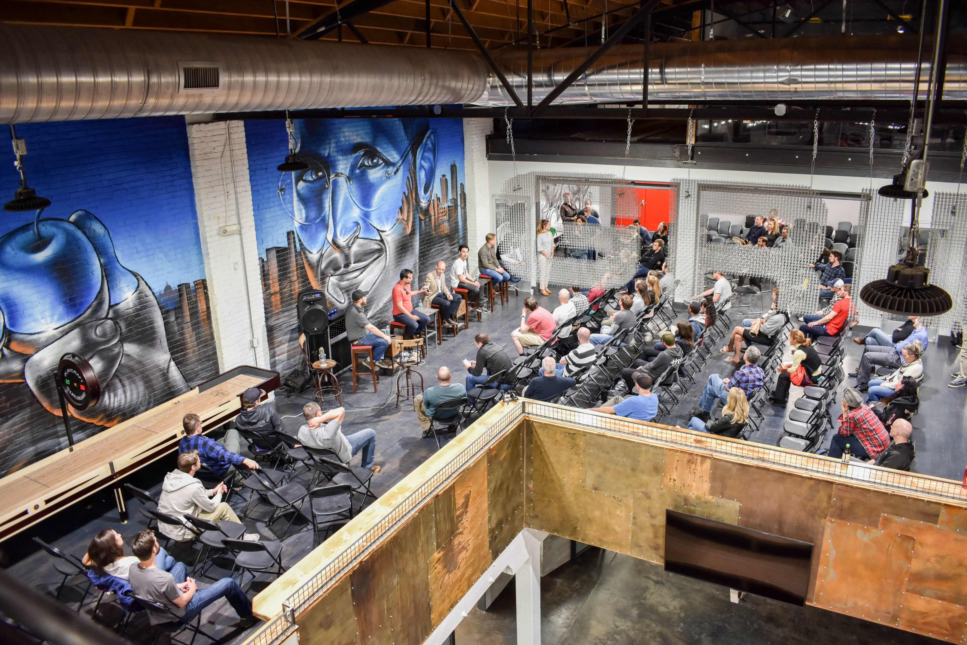 A group of speakers sits on a stage in a large, industrial-style venue with colorful murals and an audience of seated listeners.