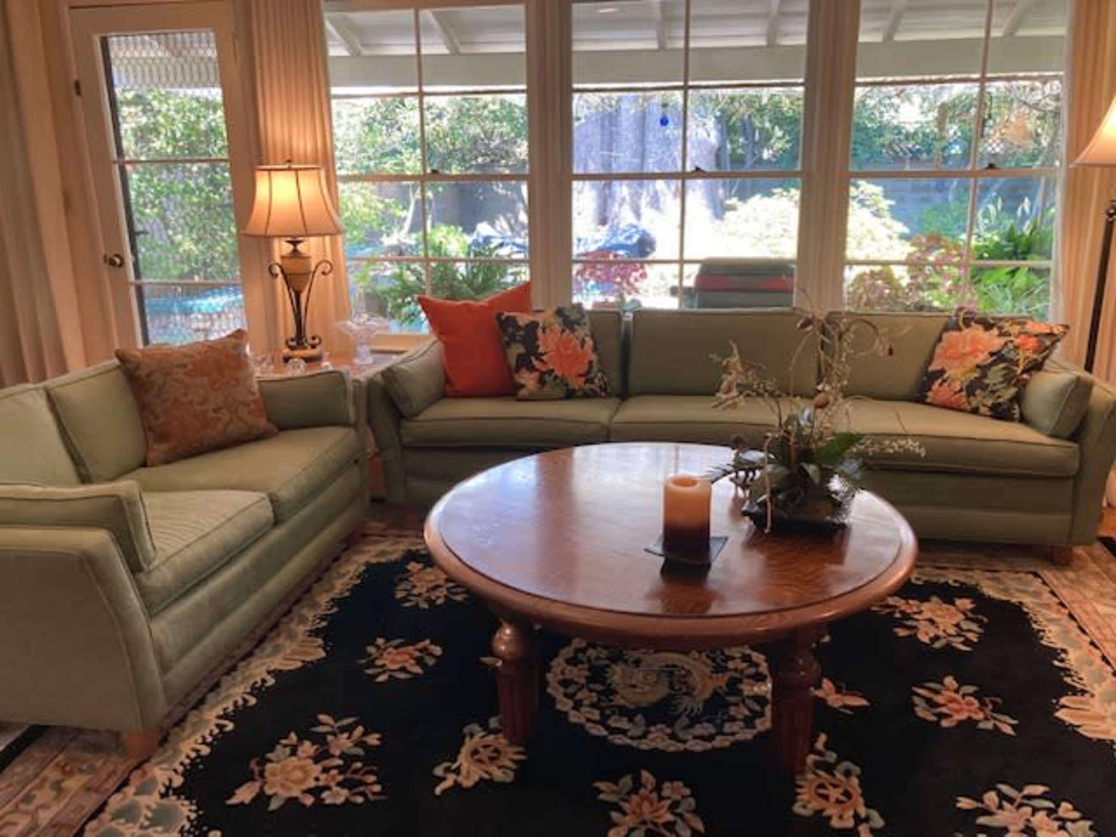 A living room features a green sectional sofa with floral pillows, a wooden coffee table, and large windows looking out onto a garden.