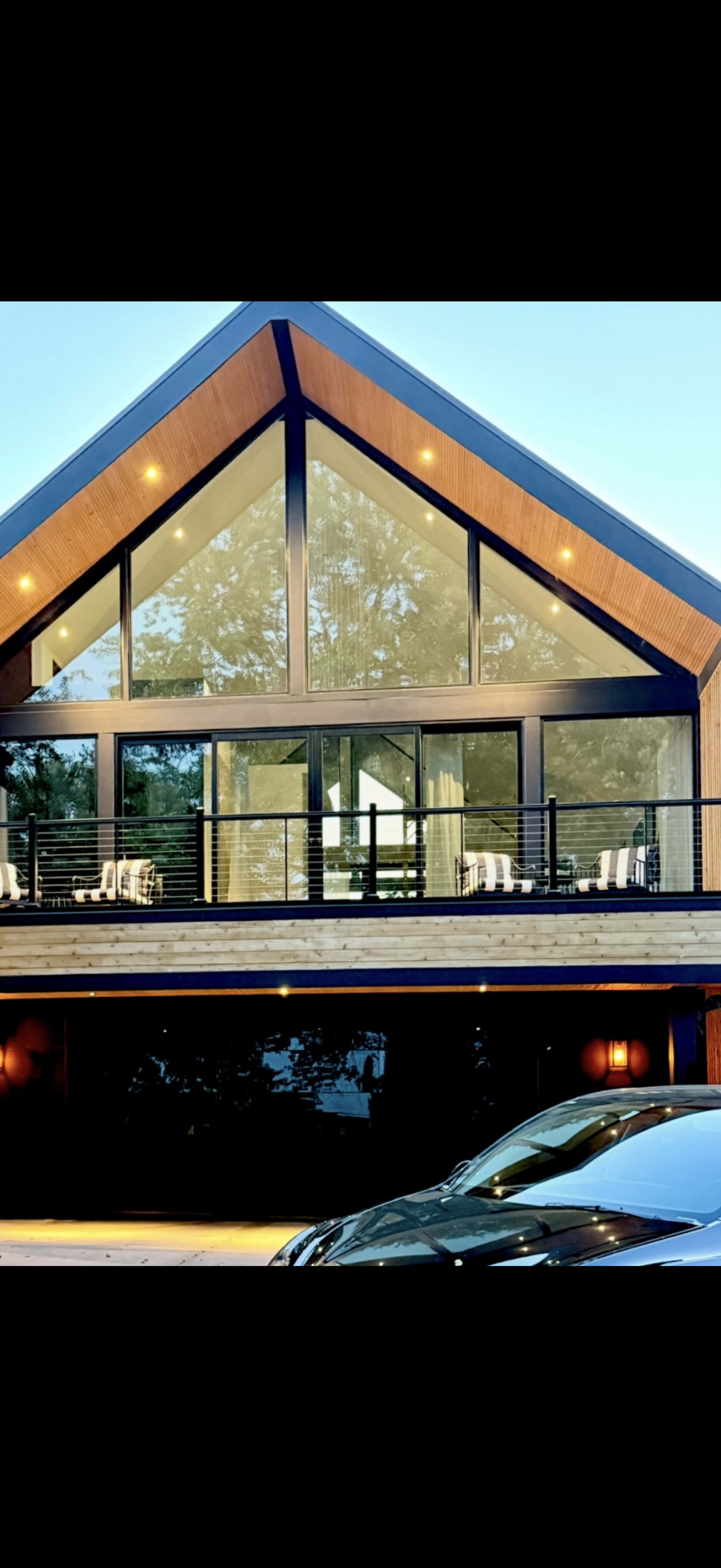 The image shows a modern house with a triangular roof, large glass windows, and a balcony overlooking a driveway with a parked car.