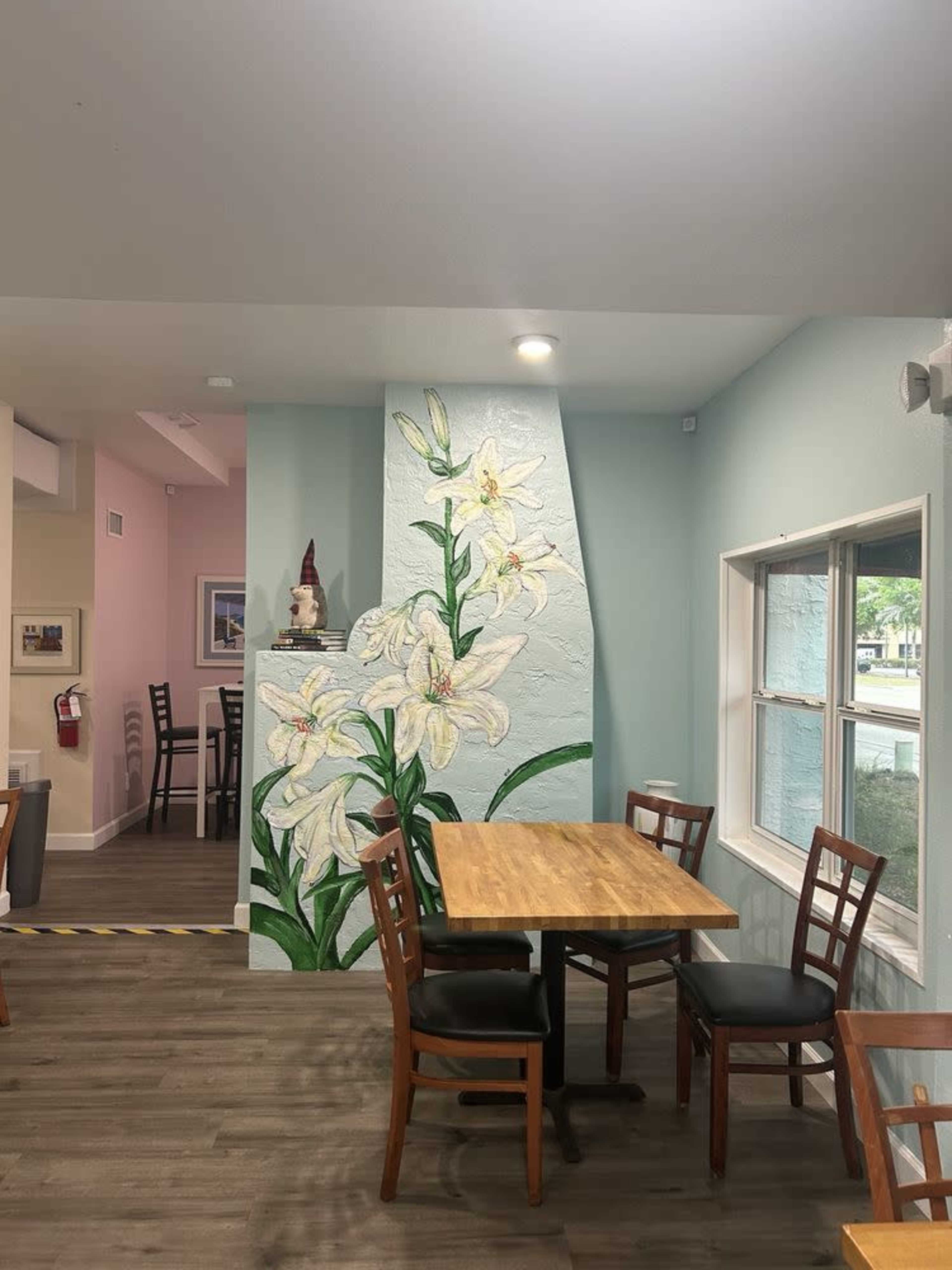 The image shows a brightly lit cafe interior featuring a wooden table with chairs beside a wall adorned with a large mural of white lilies.