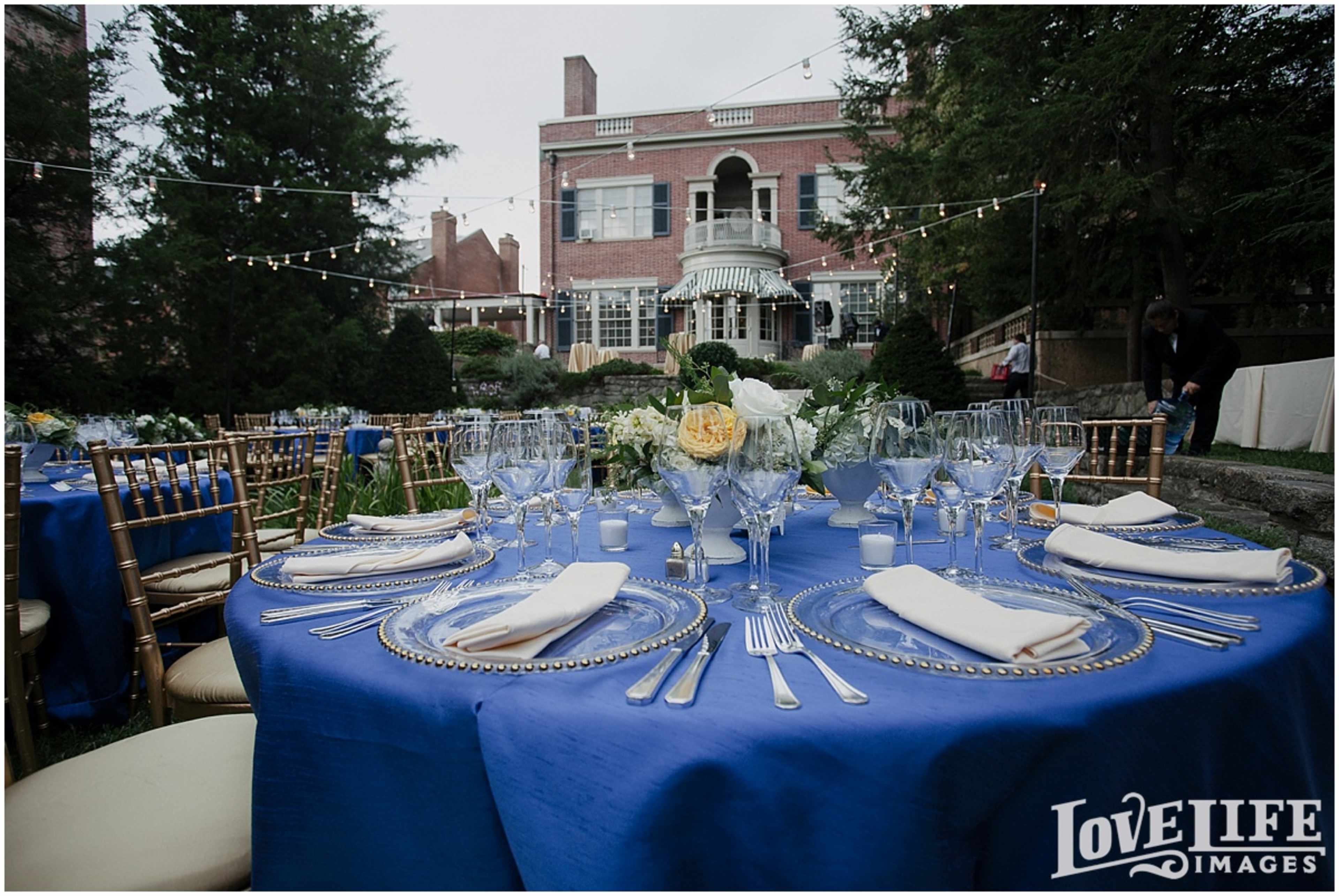 A neatly arranged outdoor dining table is set with blue tablecloths, glassware, and silverware, with a large brick building in the background.