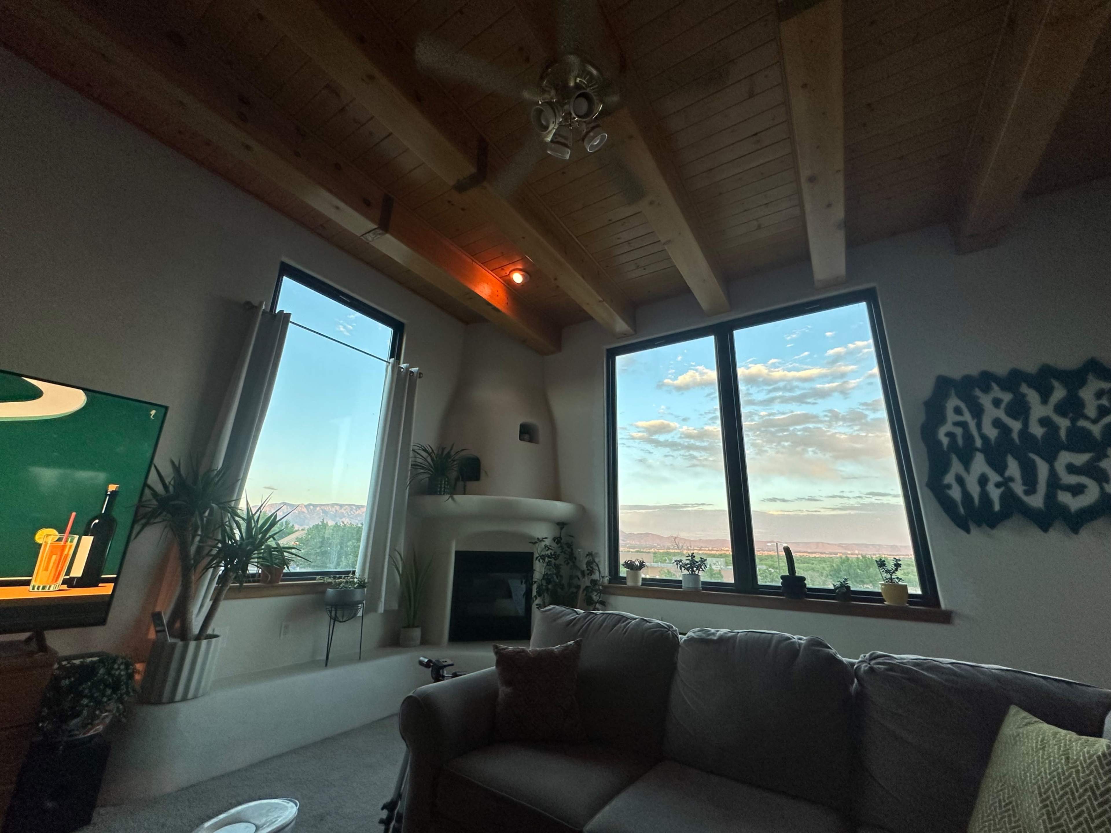 A cozy living room features large windows with a view of mountains and clouds, along with a television and plants decorating the space.