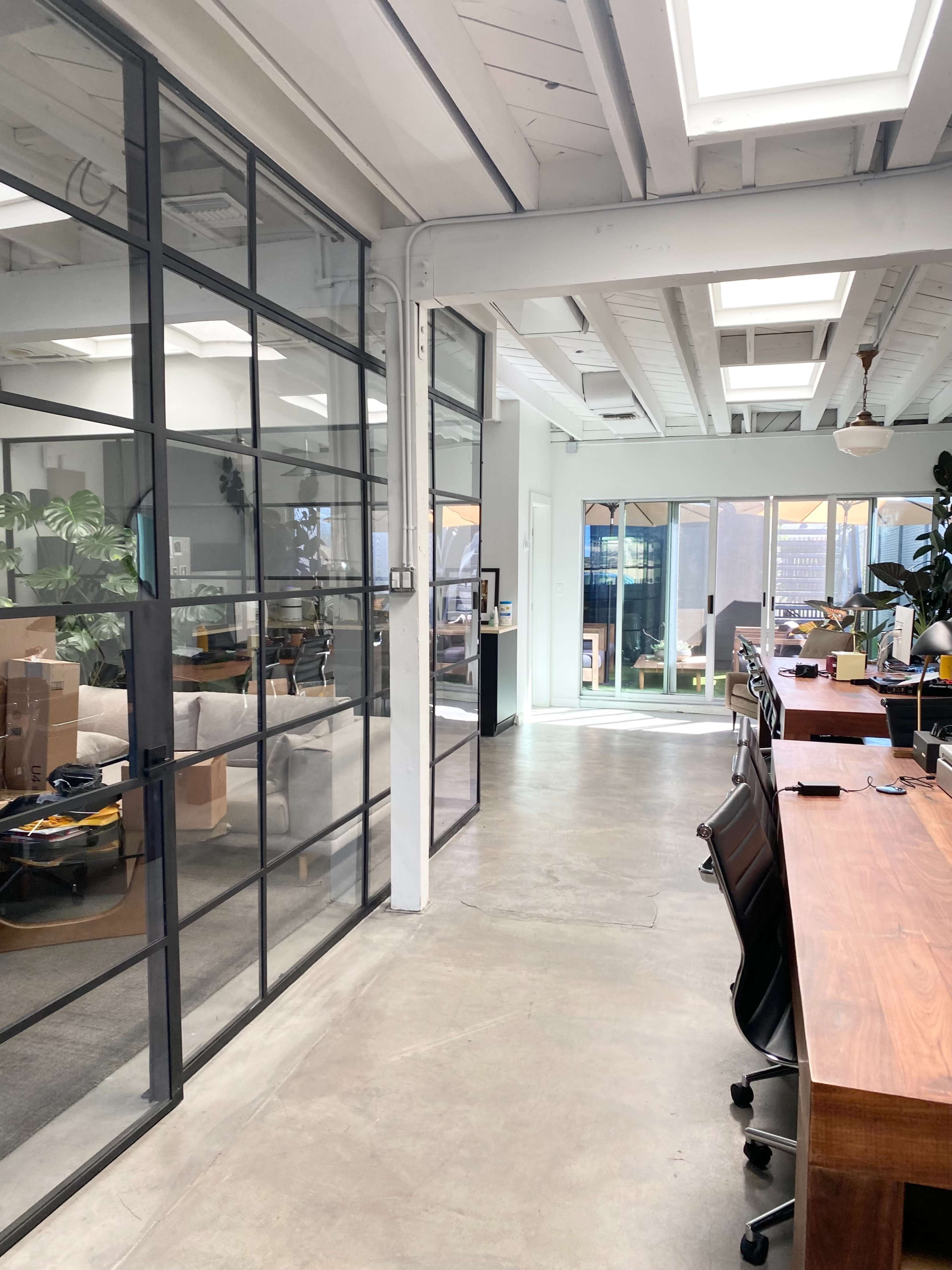 The image shows a modern office interior with large windows, a glass wall separating workspaces, and wooden desks arranged along one side.