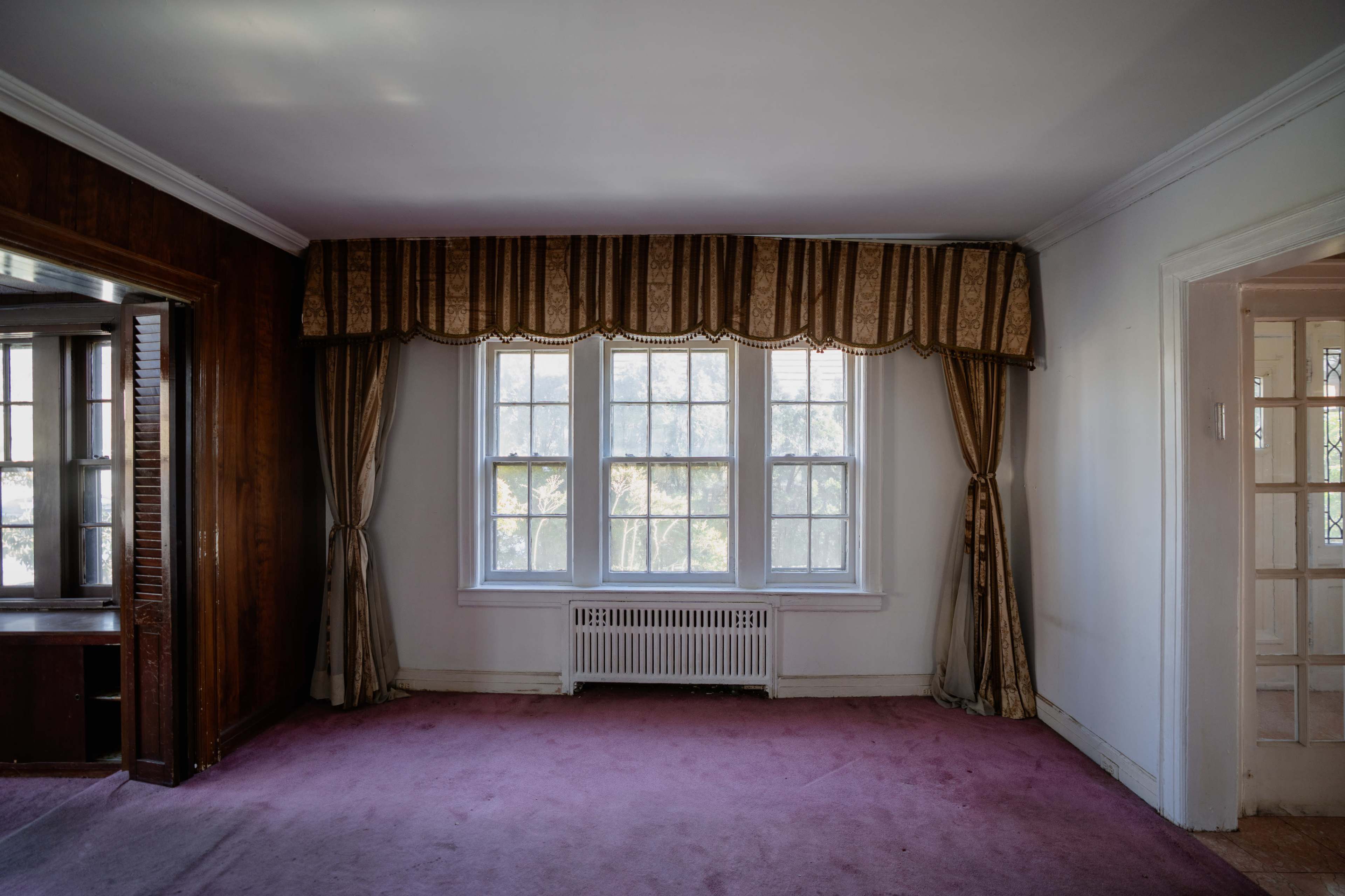 The image shows an empty room with a pink carpet, large windows draped with curtains, and a wooden wall section on one side.