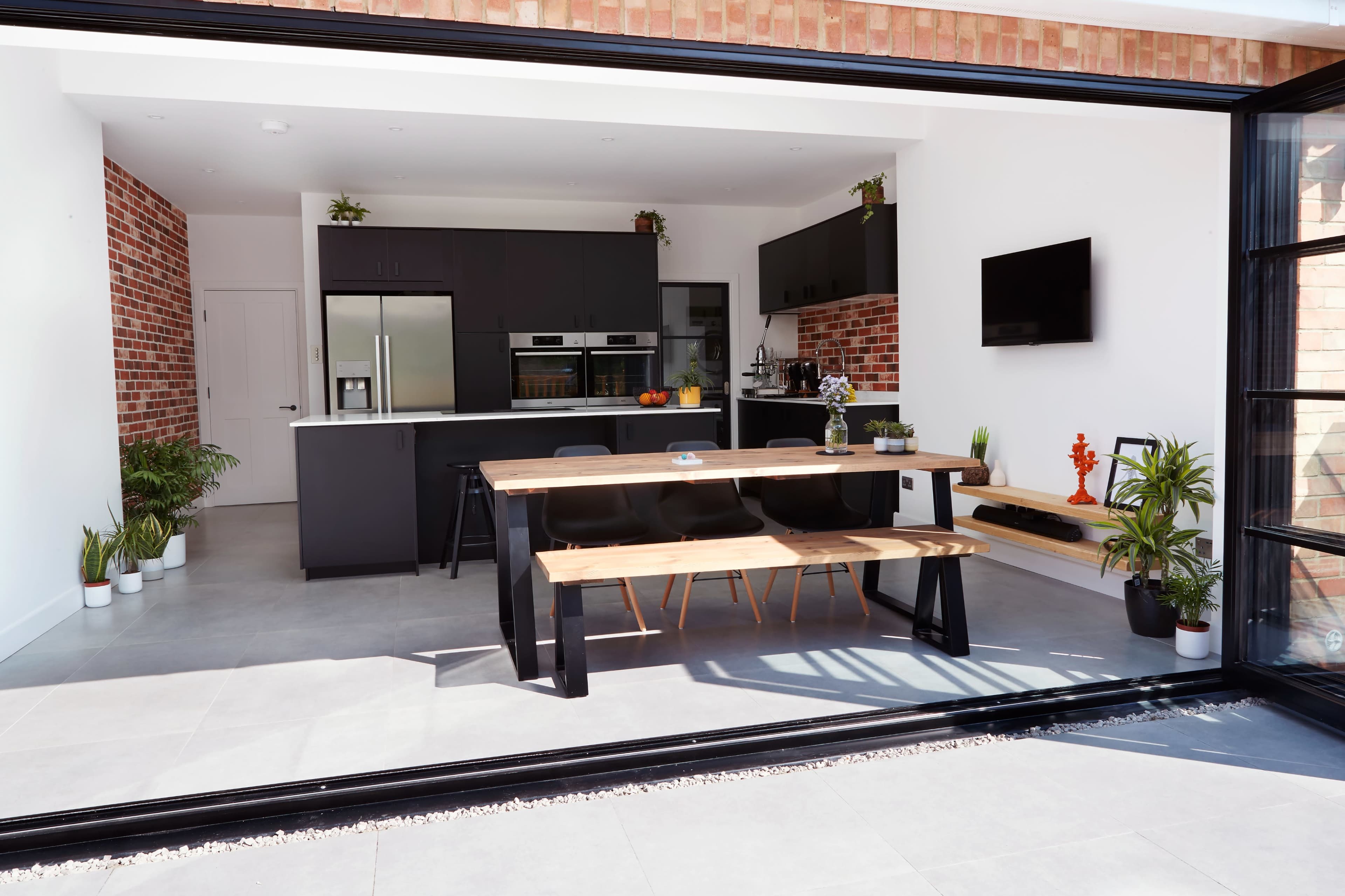 A modern kitchen and dining area features a wooden table, black cabinetry, and a large sliding glass door opening to an outdoor space.