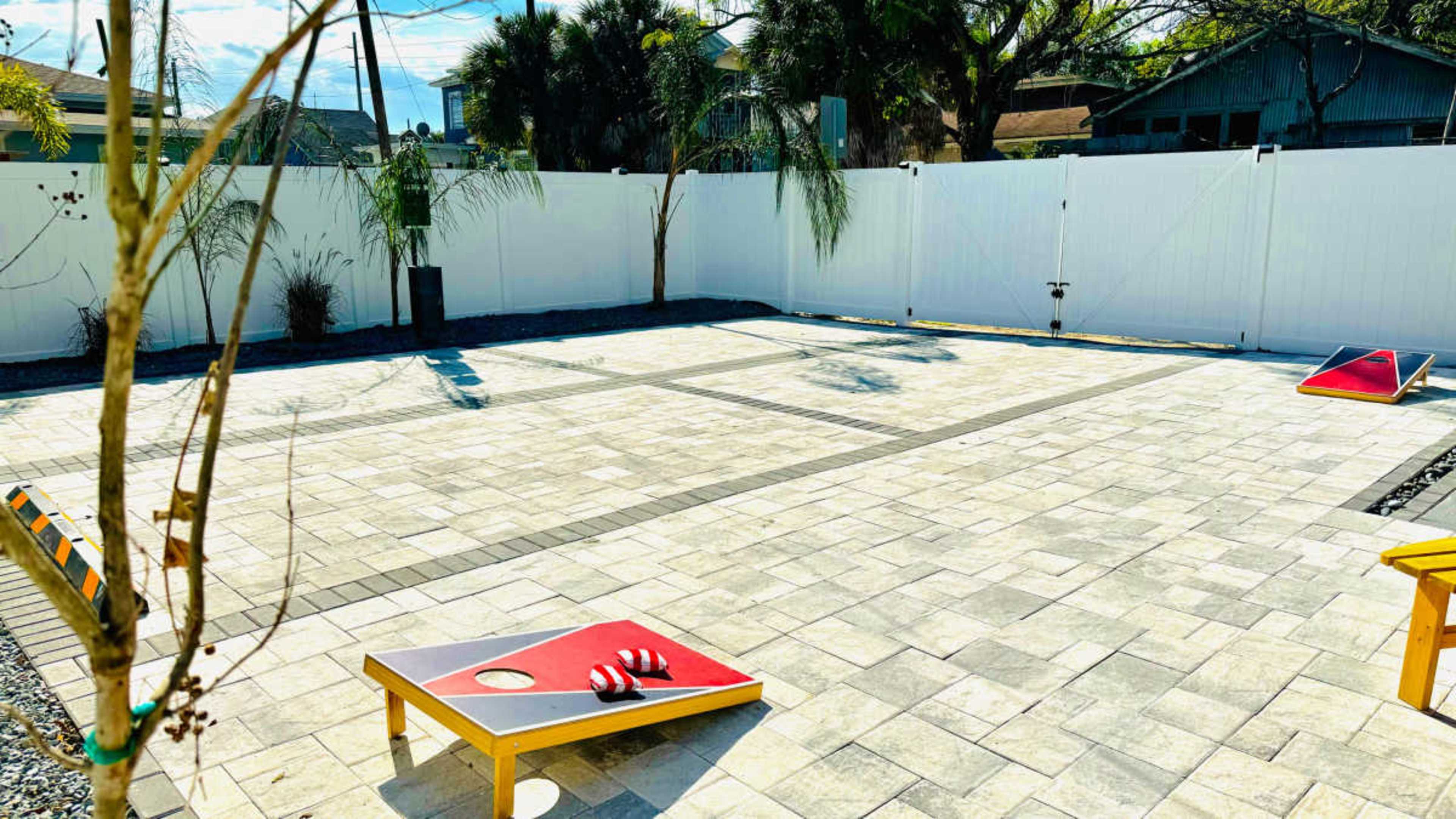 The image shows a patio area with a paved stone surface, bordered by a white fence, featuring four cornhole boards and some landscaping elements.