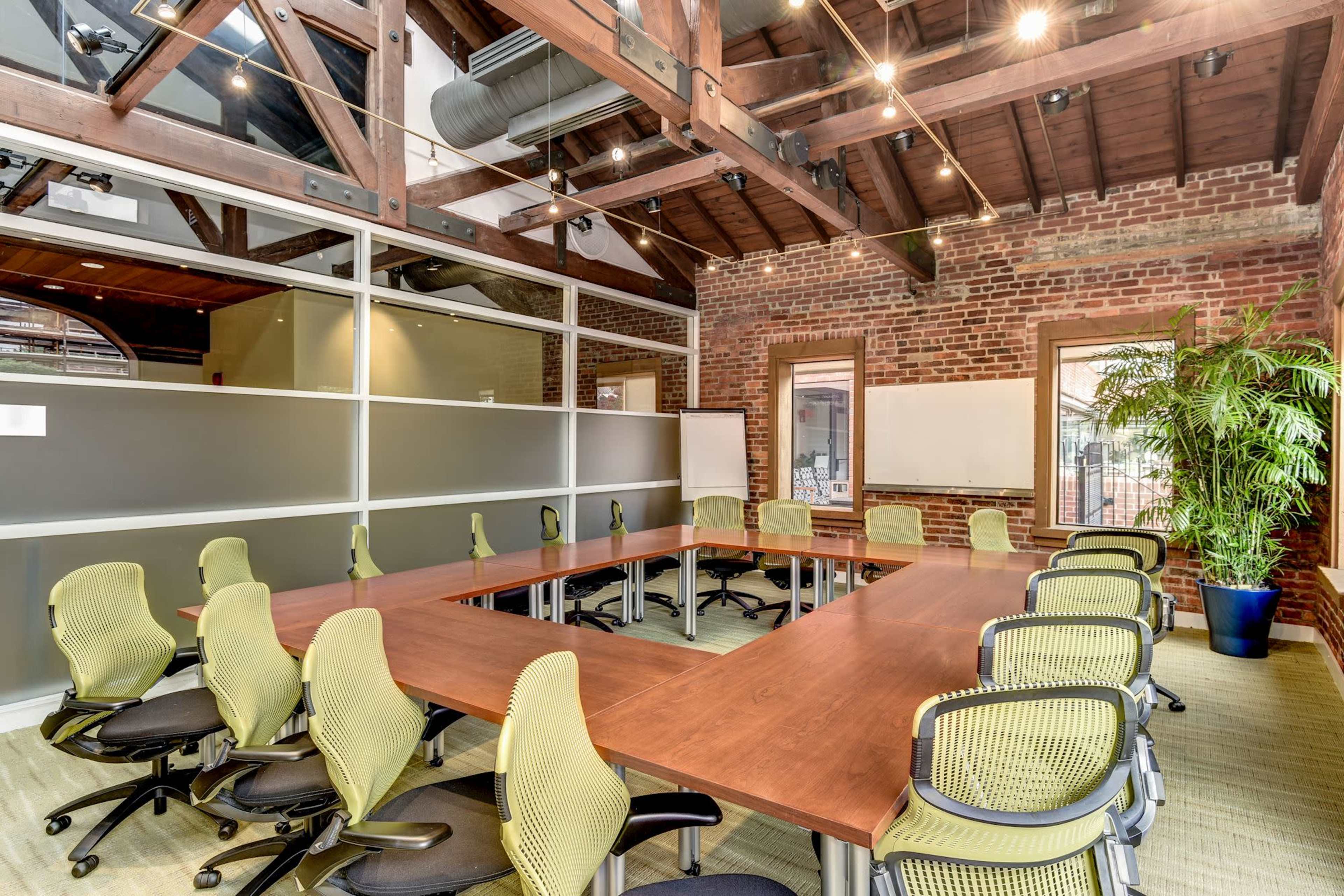 A spacious conference room with a large wooden table surrounded by green office chairs, exposed brick walls, and large windows.