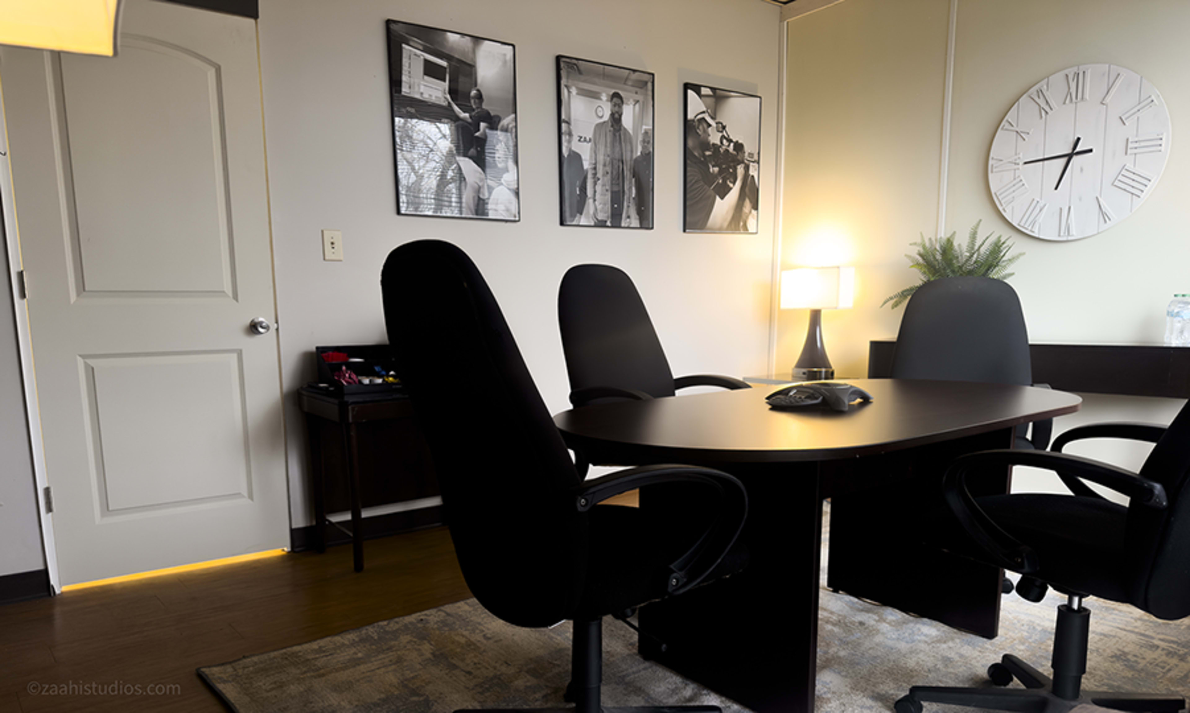 The image shows a conference room with a round table, four black chairs, a clock on the wall, and framed black-and-white photographs.