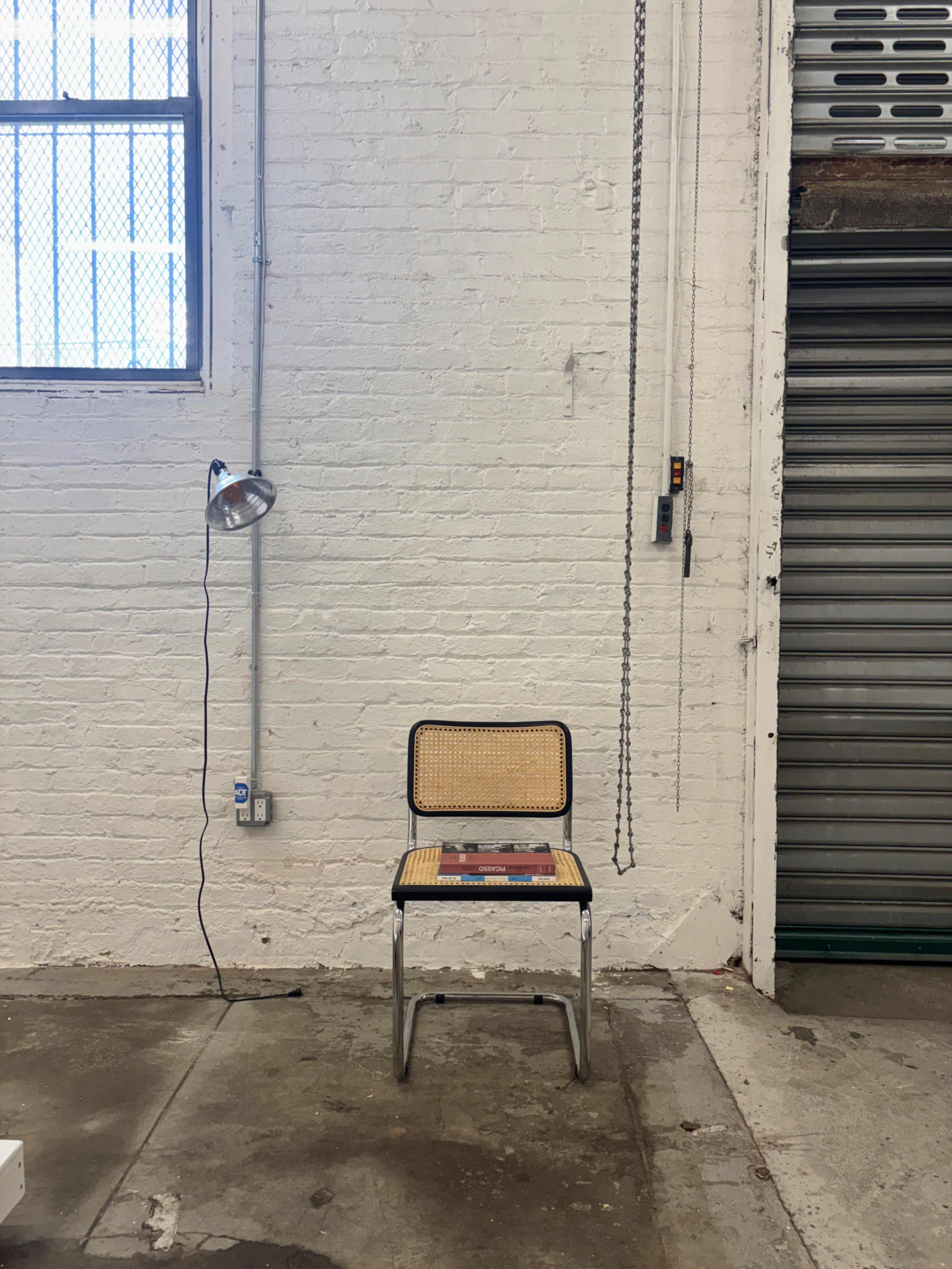 A metal chair with a cane seat is positioned against a white brick wall, illuminated by a lamp hanging nearby.