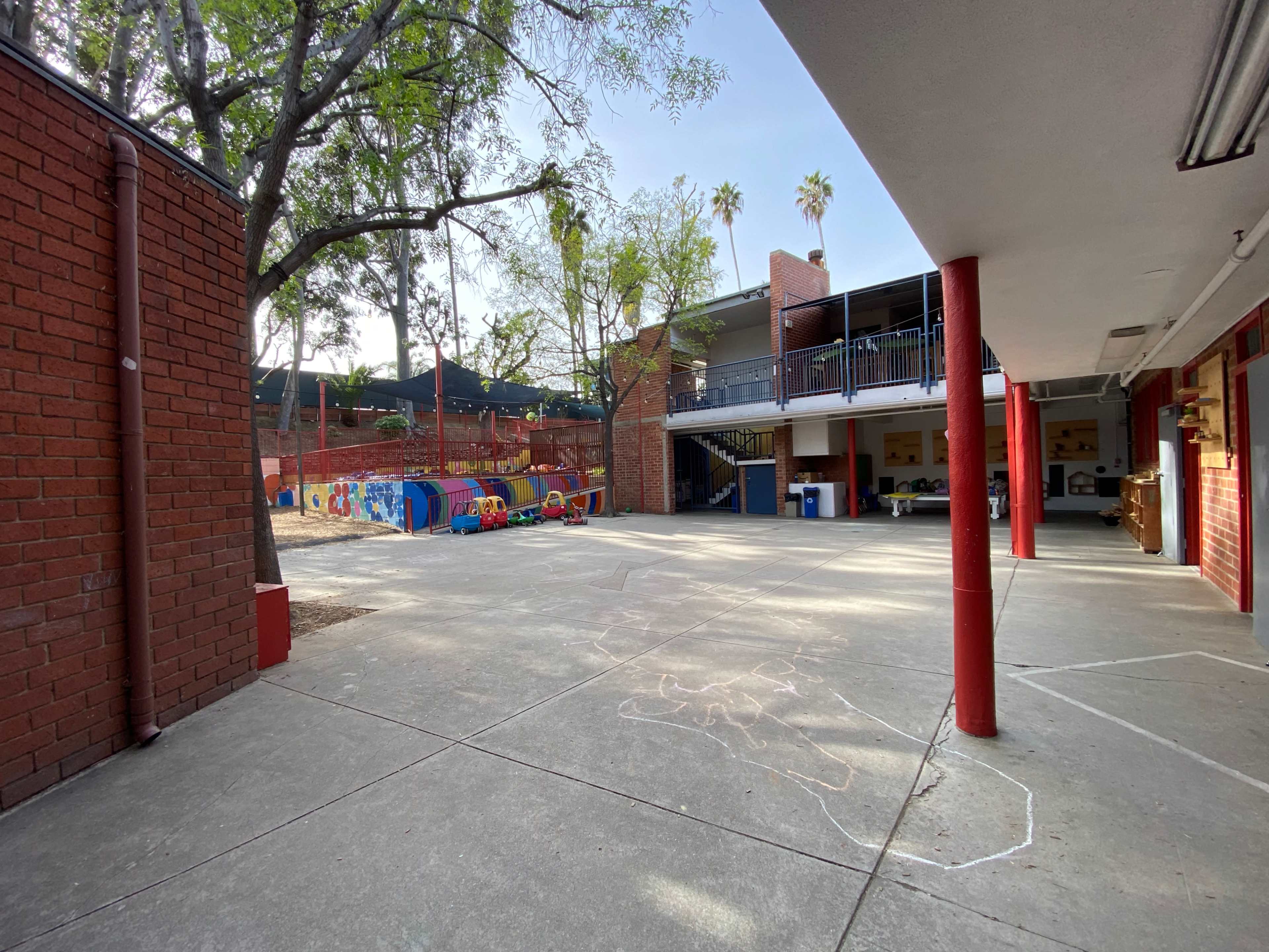 A school playground area with colorful play equipment, adjacent to a building with red columns and a balcony.