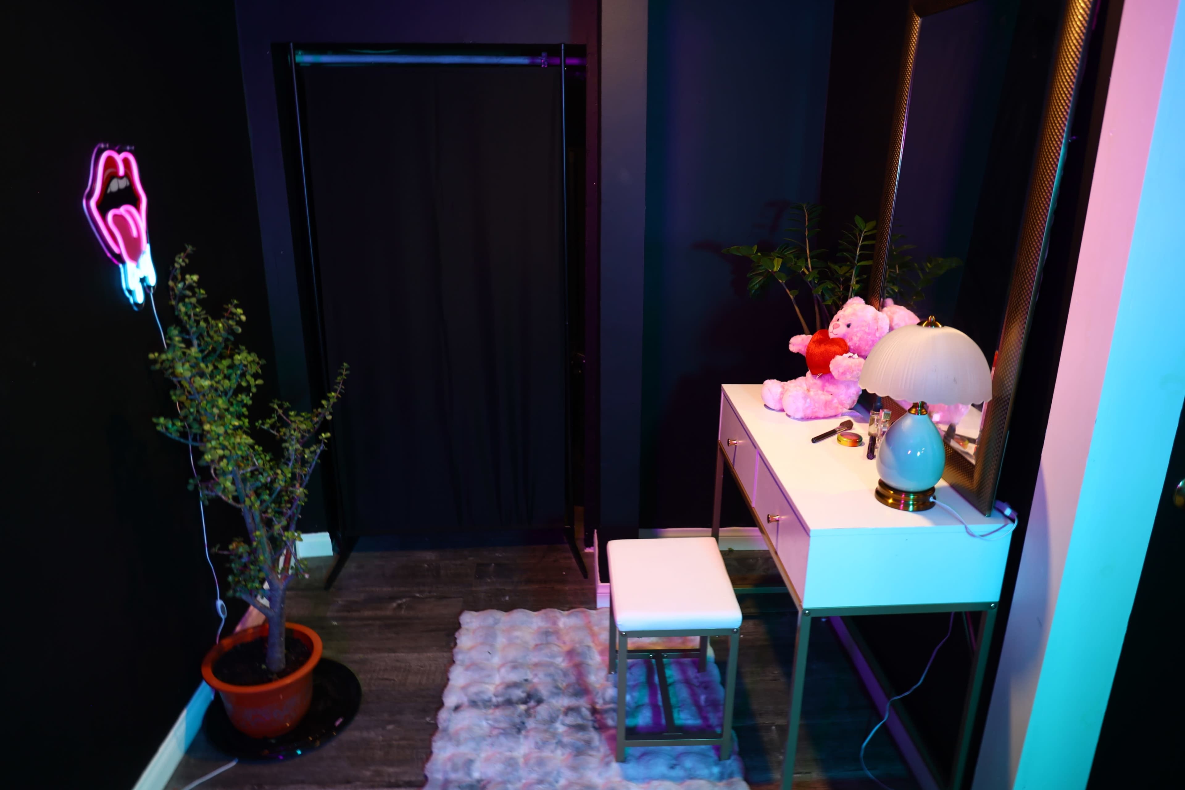 The image shows a small, modern vanity area with a white table, a stool, a lamp, a decorative plush toy, and a potted plant, all set against a dark wall.
