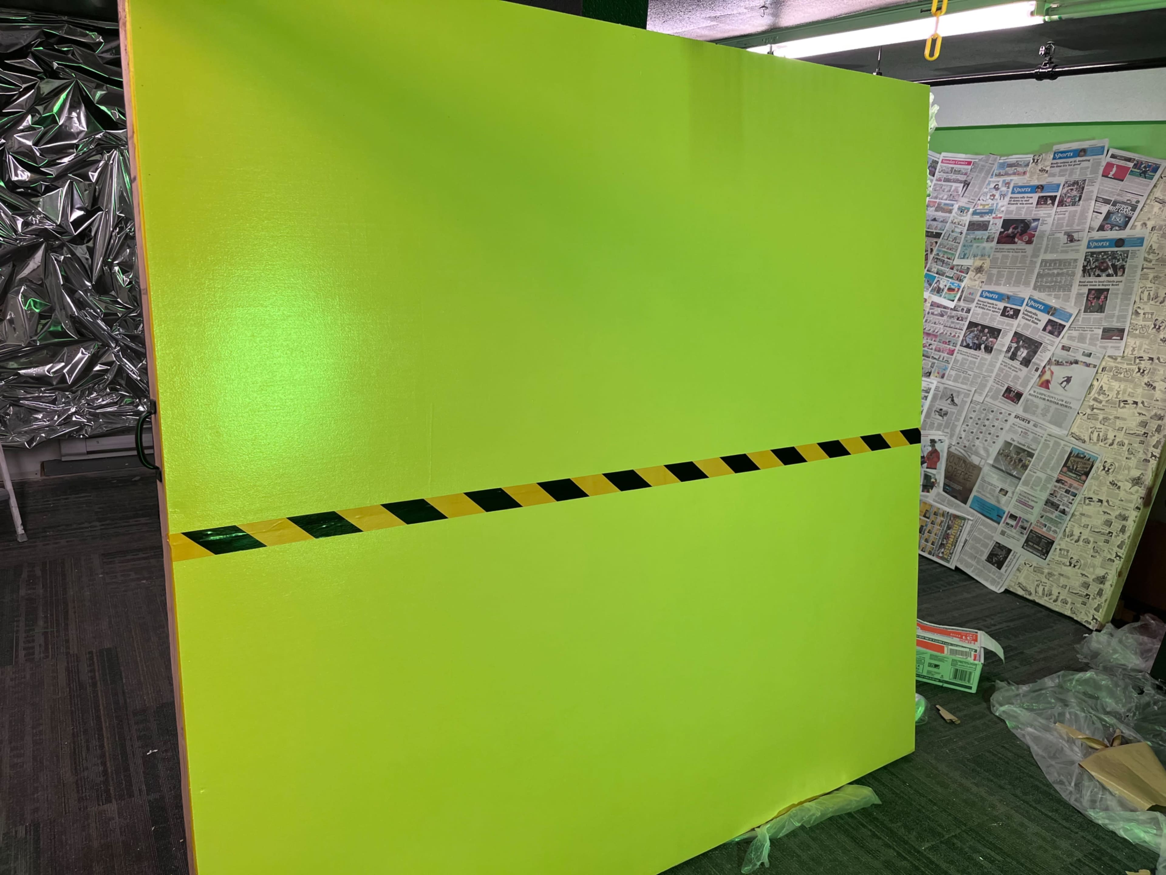 A large green wall with a black and yellow striped caution tape running horizontally across it stands in an indoor space, adjacent to a backdrop of newspaper clippings.