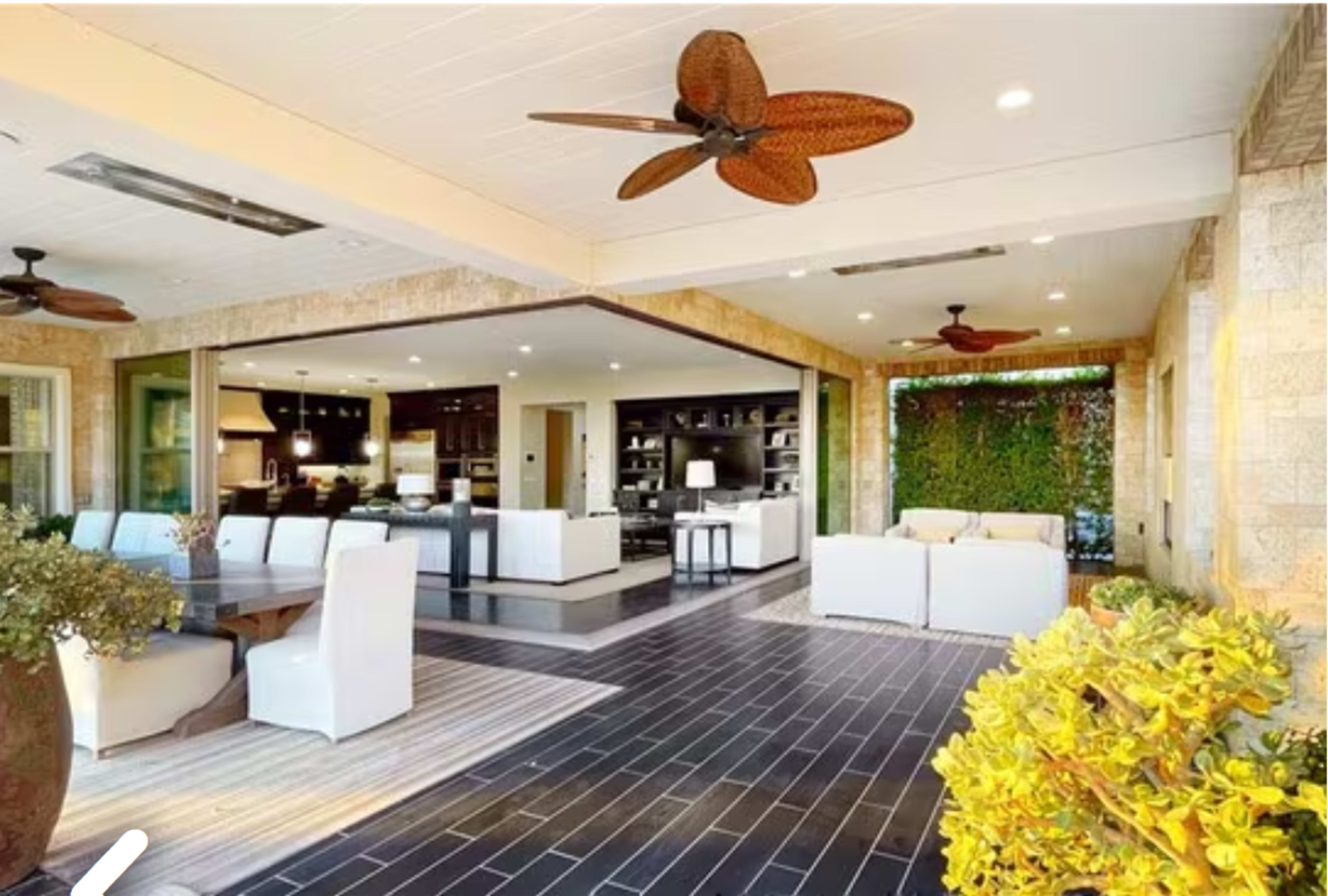 The image shows a modern outdoor living area with a dining table and seating, bordered by large glass doors that open to an indoor space featuring a kitchen and shelves.