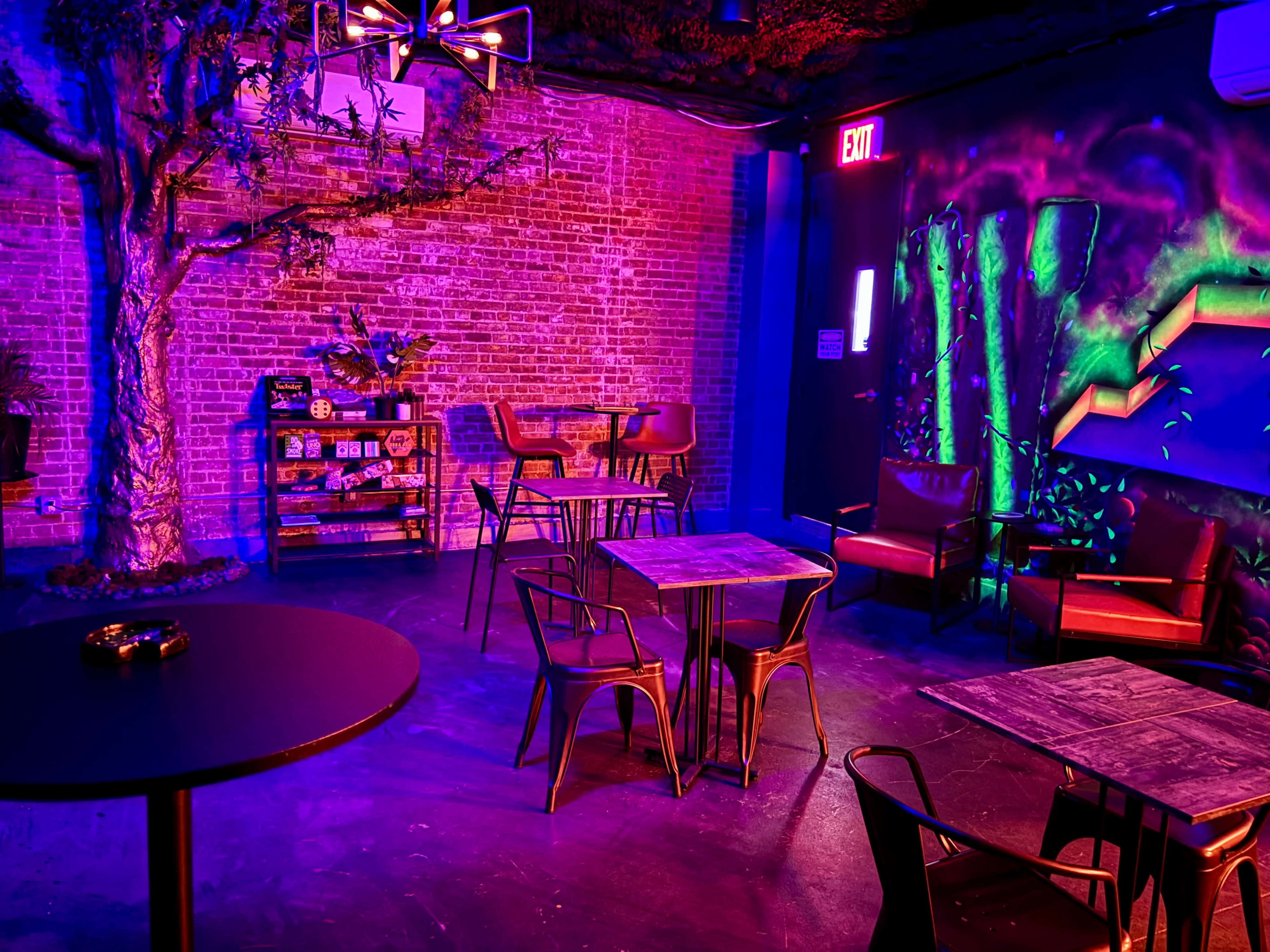 The scene depicts a dimly lit lounge area with colorful neon lighting, featuring tables and chairs arranged around a brick wall, along with a shelf displaying various items.