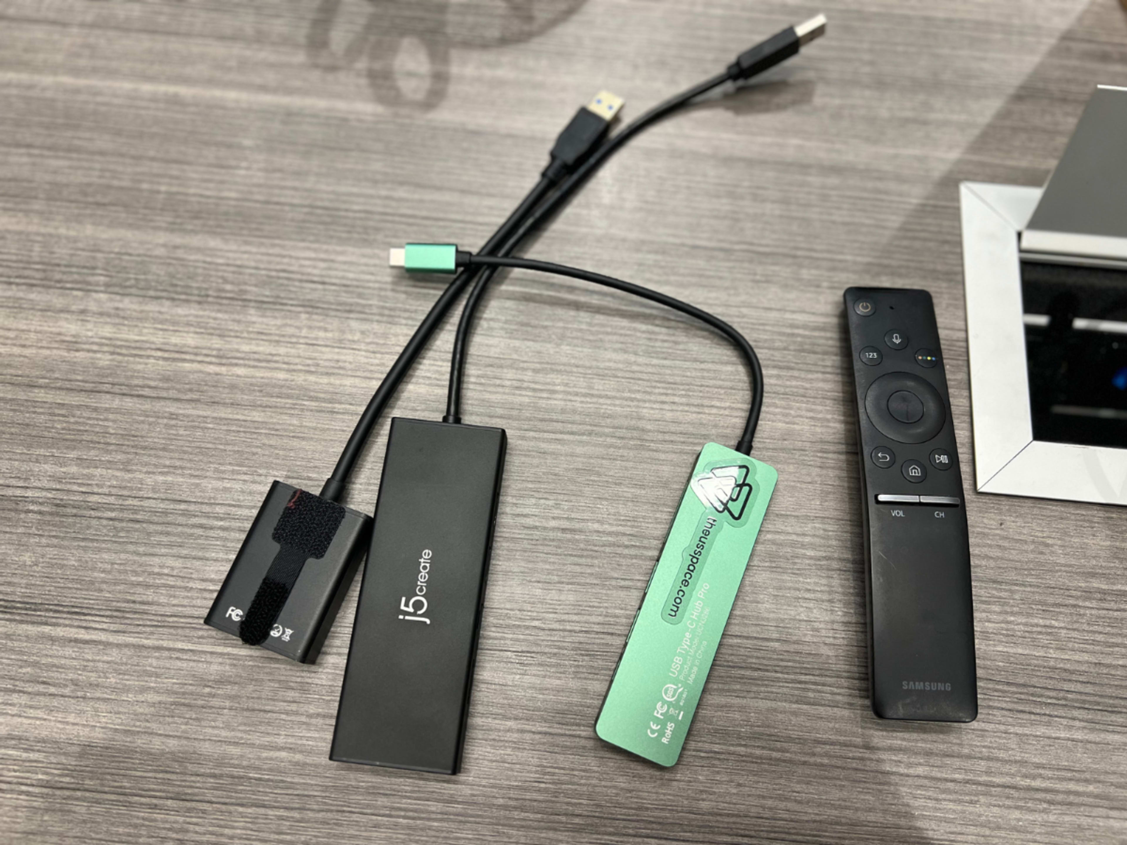 A j5create USB hub connected to a laptop, accompanied by a remote control and a power bank on a wooden surface.