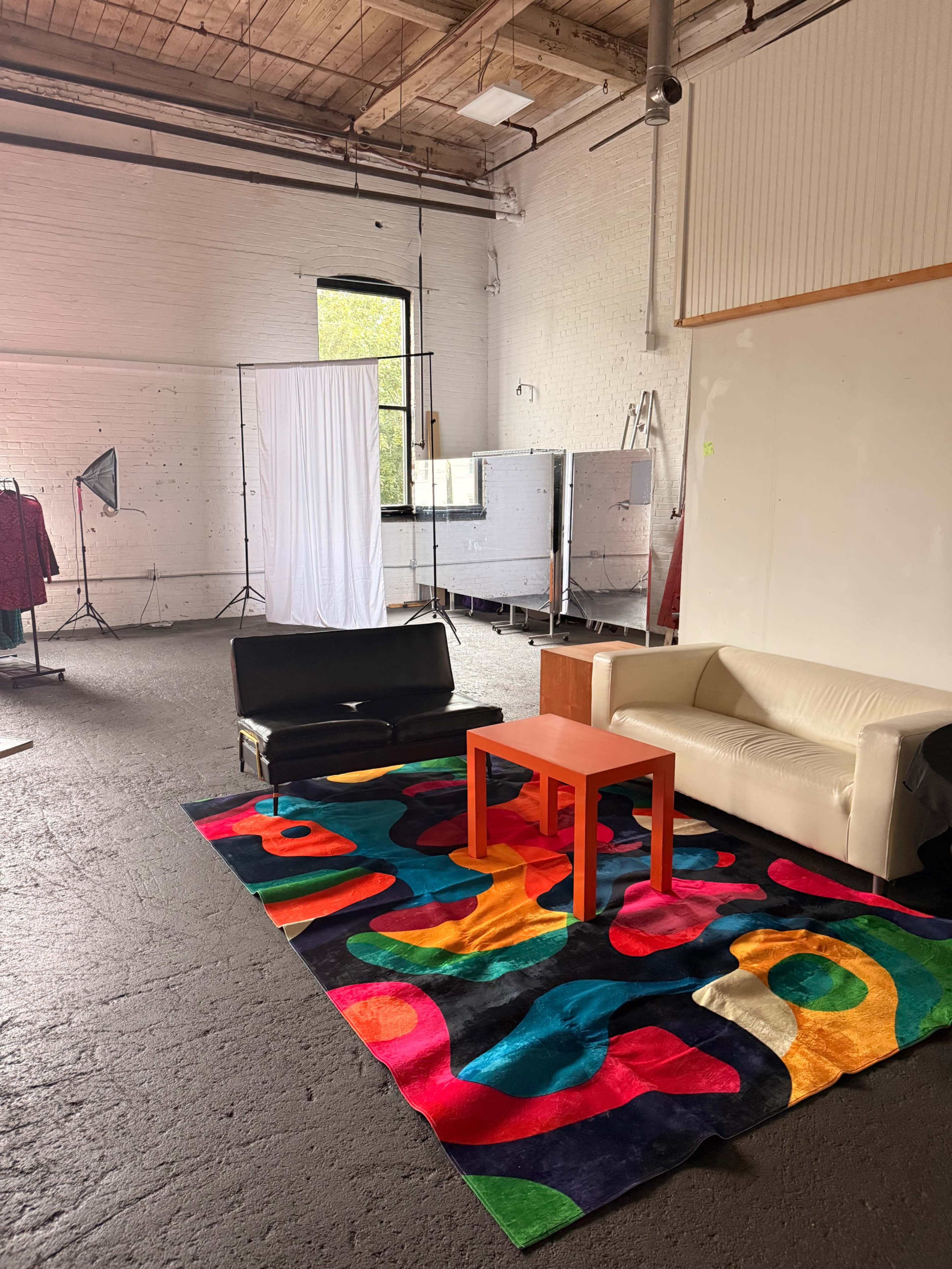 The image shows a spacious, well-lit room with a colorful rug, a black couch, a white sofa, and an orange coffee table, alongside photography equipment in the background.