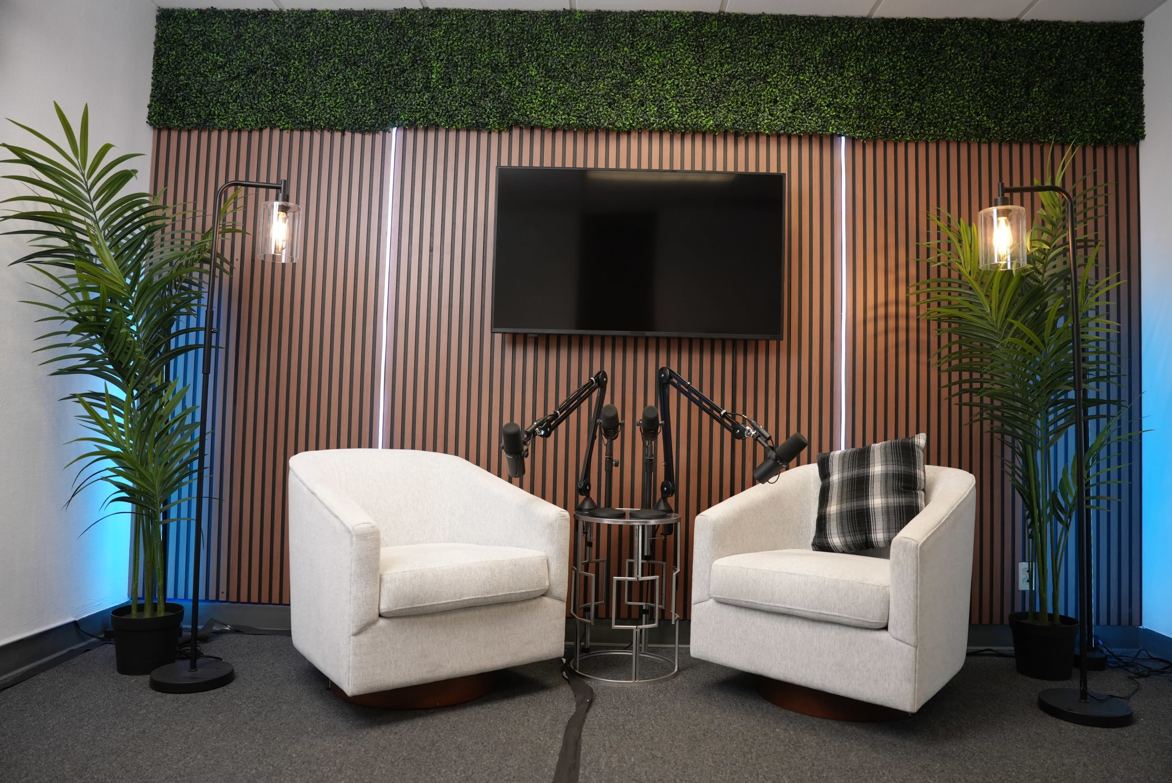 The image shows a modern interior setup with two white armchairs, a small table, and a wall-mounted TV, all surrounded by decorative plants and bamboo-like paneling.