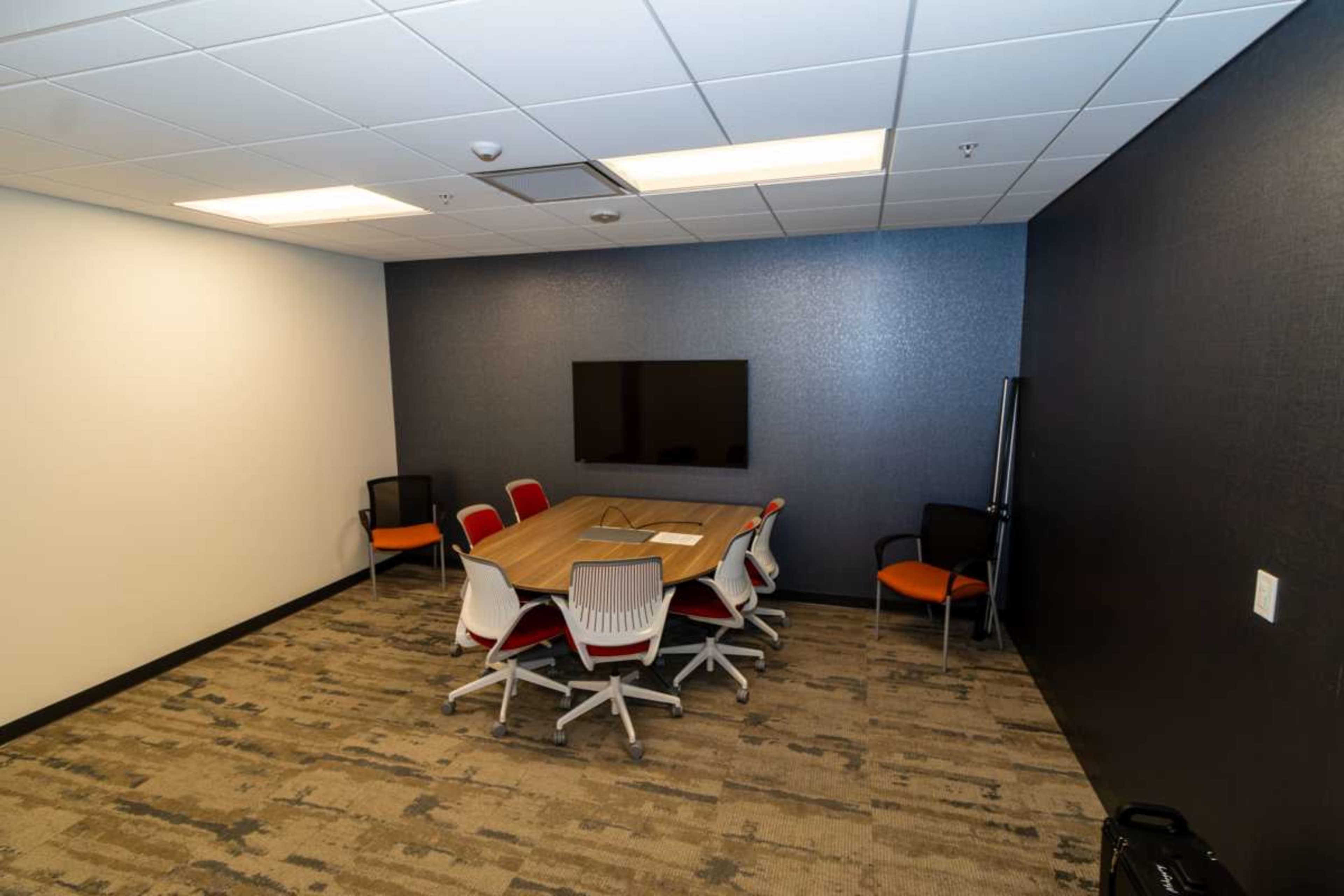 The image shows a modern conference room with a large table surrounded by eight chairs and a wall-mounted television.