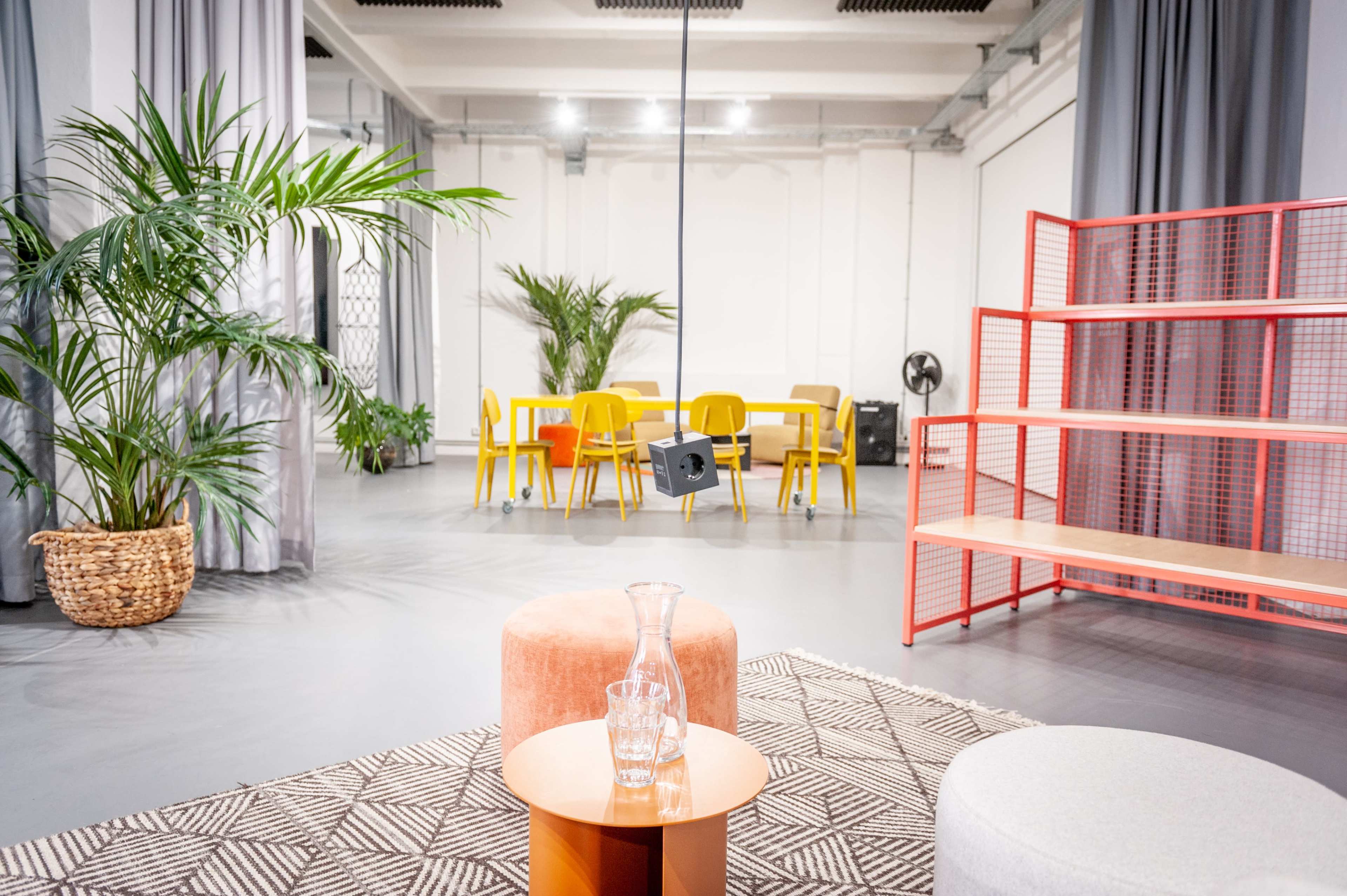 A modern interior space with a seating area featuring round stools, a small table, a small plant, and a dining area in the background with yellow chairs and a red shelving unit.