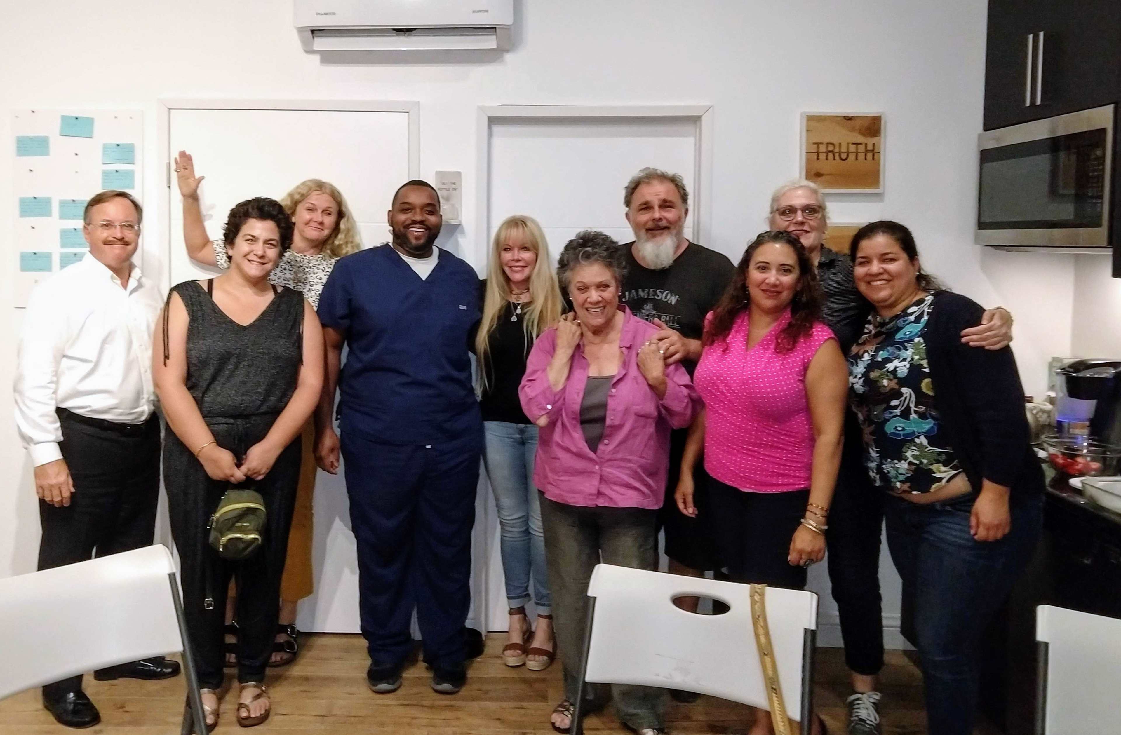 A group of ten people poses together in a well-lit room with kitchen facilities and a sign that reads "TRUTH."