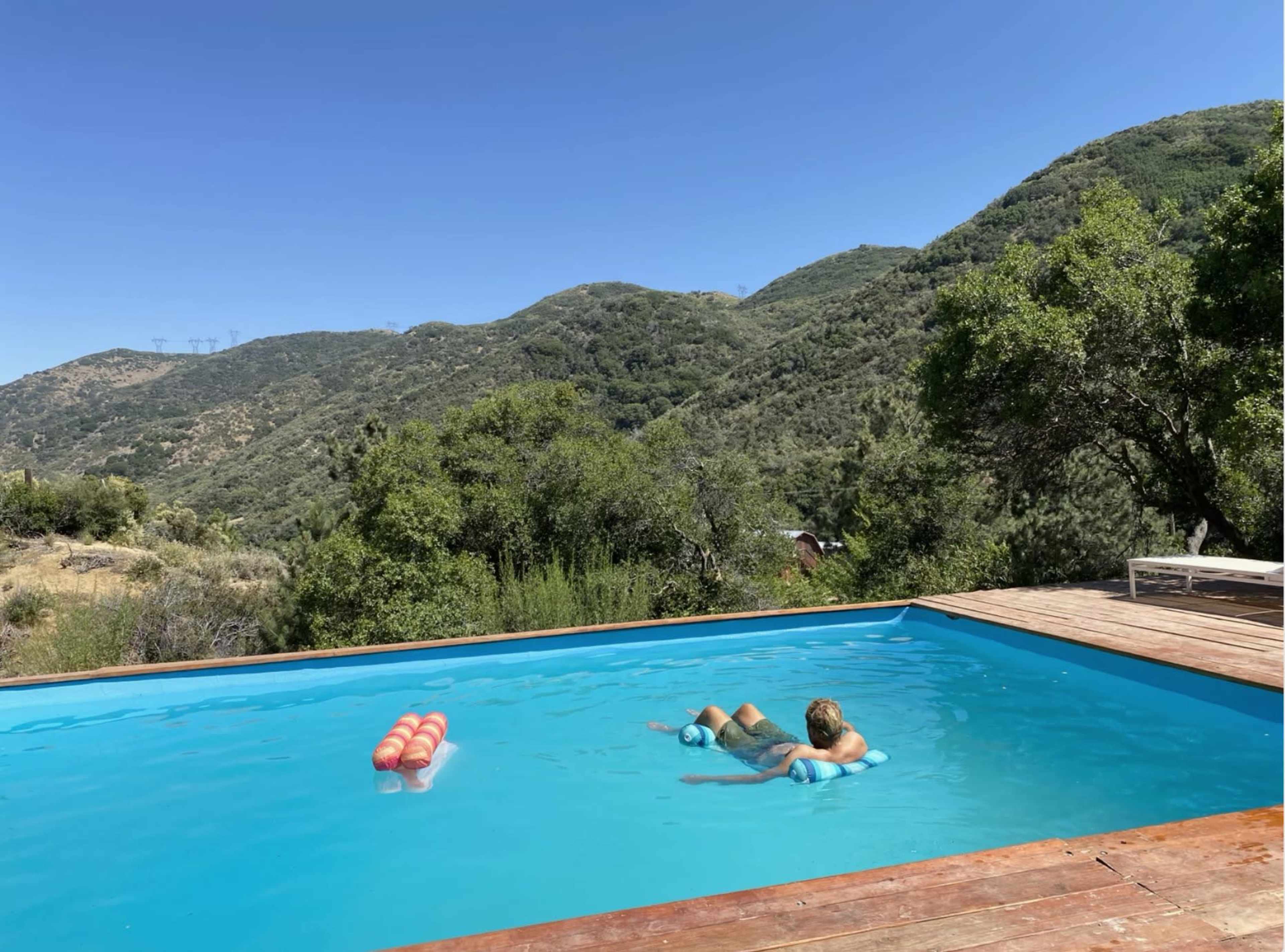 A person relaxes in a blue swimming pool with mountains in the background.
